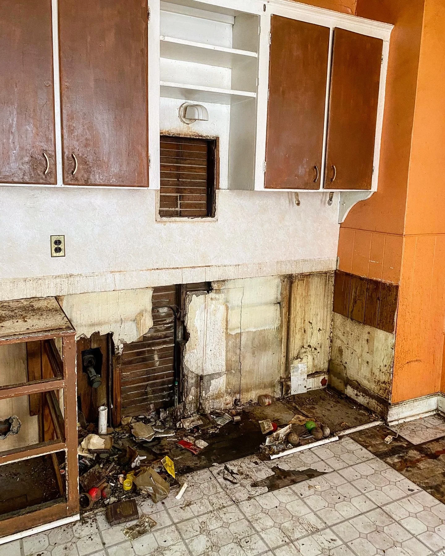 1915 kitchen before it's renovated with damaged walls, vinyl floors, and brown cabinets that are partially demolished showing grease and trash.