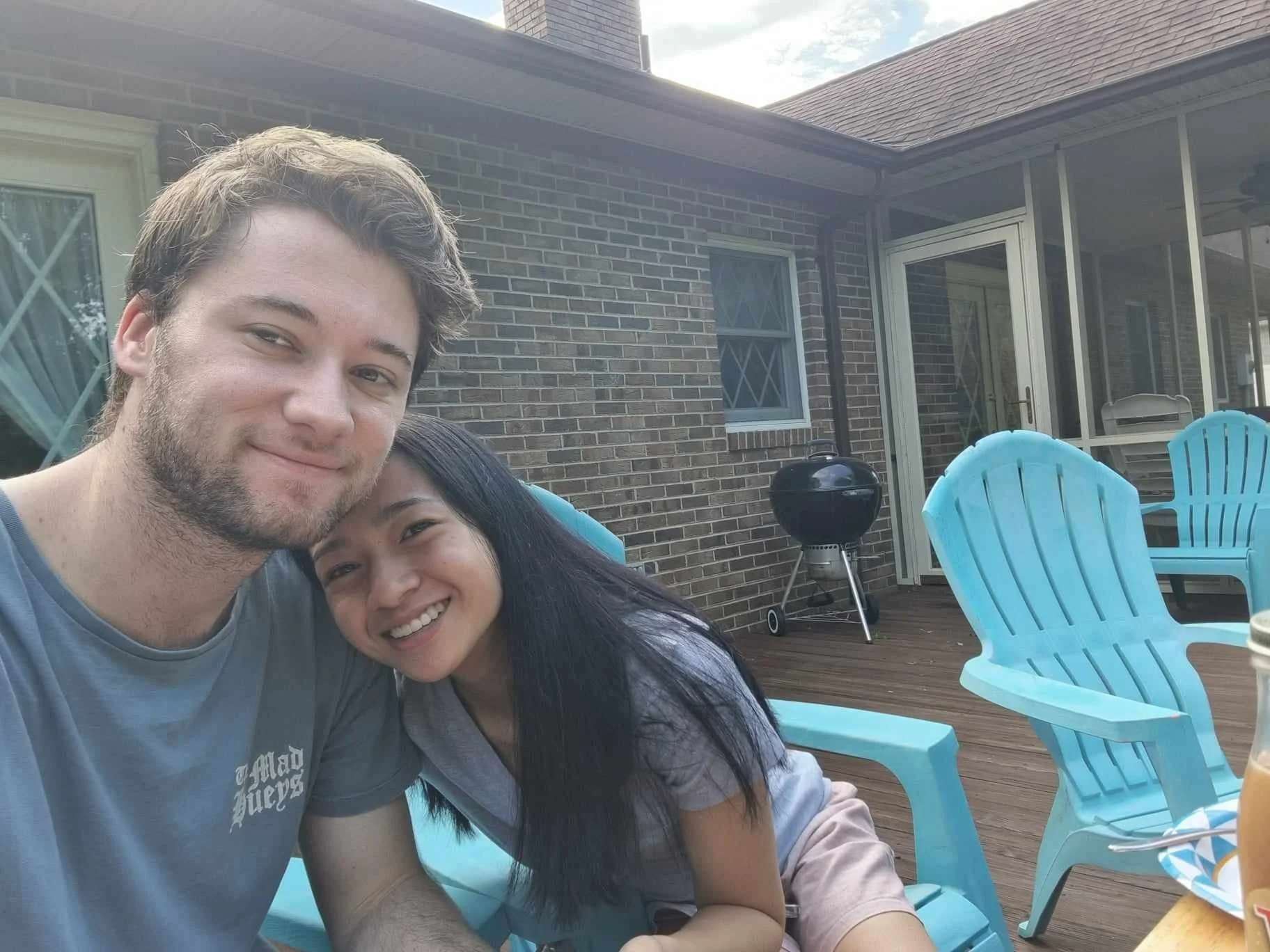 A young man and woman smiling and sitting close together on a patio with blue chairs, a black grill, and a brick house in the background.