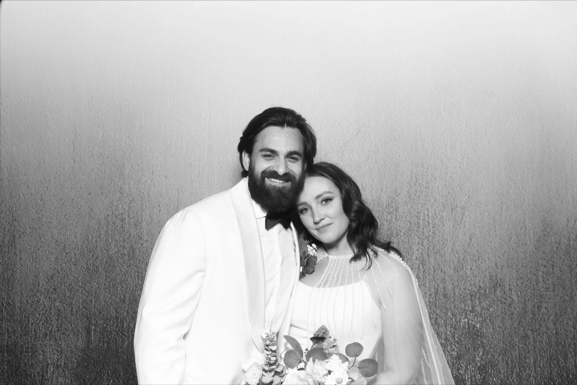 Black and white photo of a happy bride and groom on their wedding day, standing closely with the groom dressed in a tuxedo and the bride holding a bouquet.