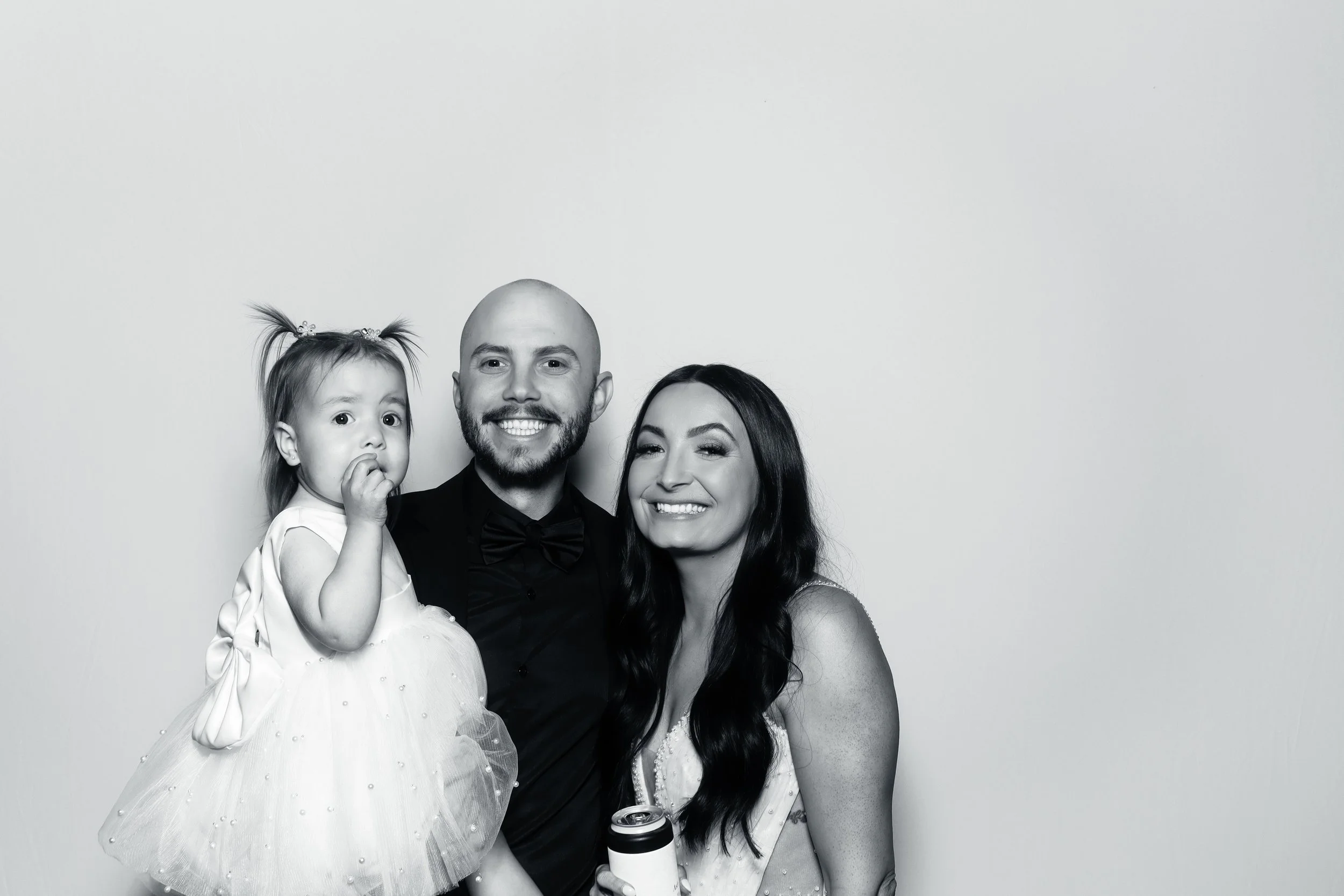 Black and white photo of a happy family of three posing together against a plain background. The man is holding a young girl and the woman is smiling with a can in her hand.