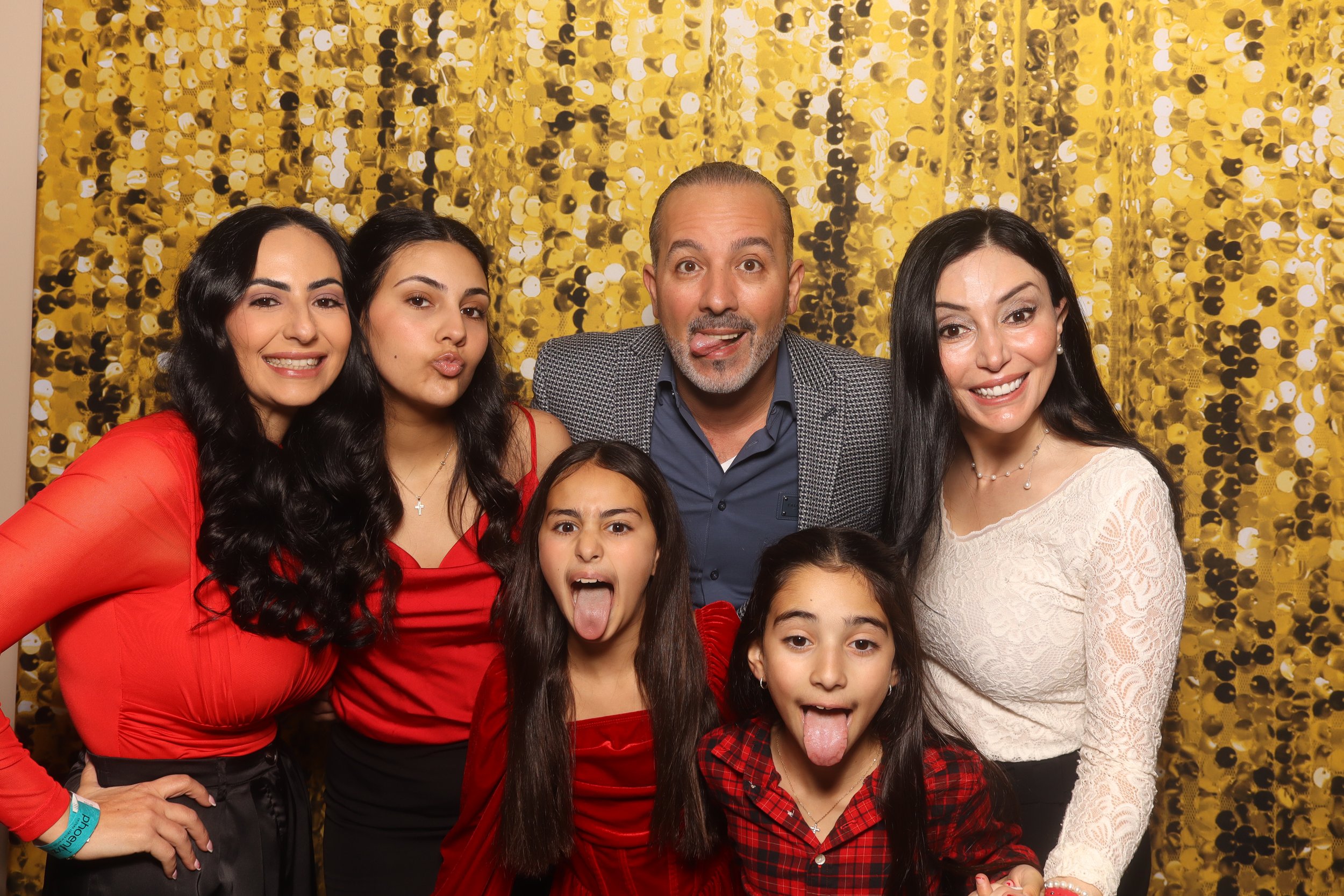 Group of six people, including three women, one man, and two young girls, posing together in front of a gold sequin backdrop, smiling and making funny faces.