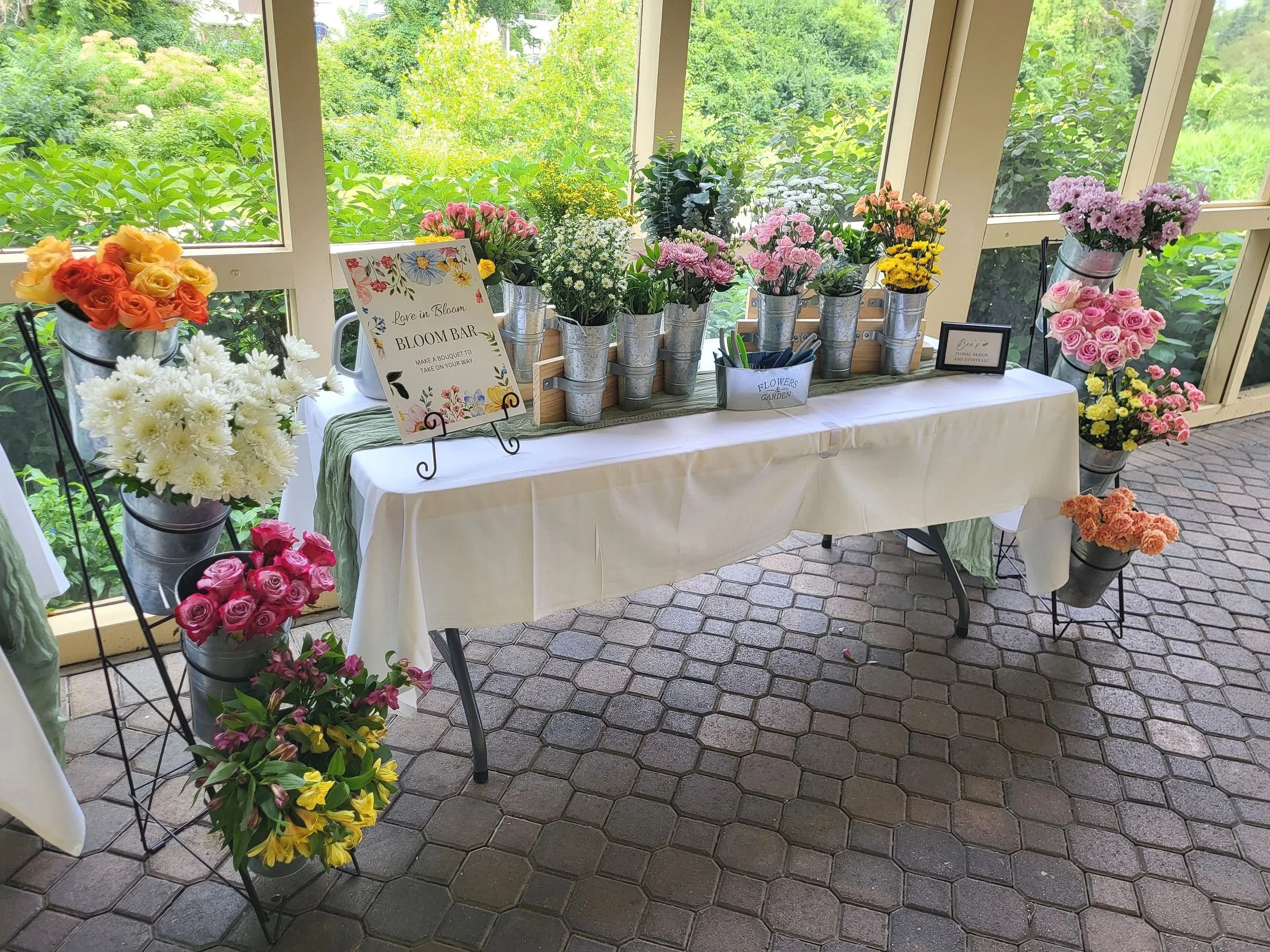 Display of various colorful flowers arranged in buckets and on tables, with signs indicating a flower shop or event setup, inside a greenhouse or conservatory with glass windows and lush greenery outside.