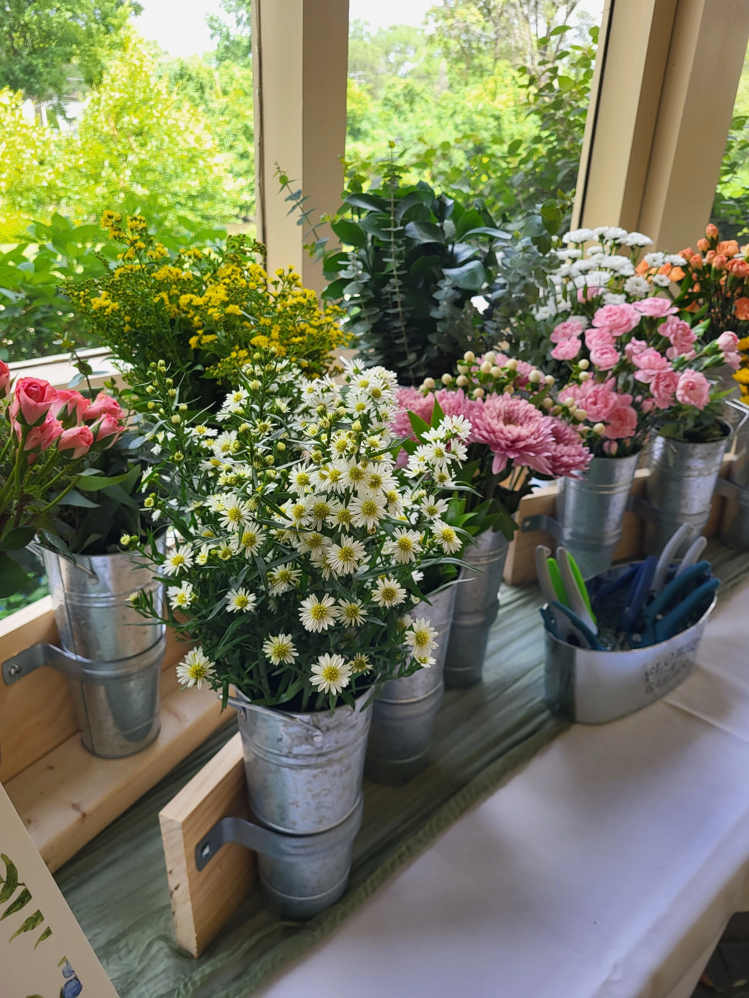 Various colorful flowers arranged in metal buckets on a wooden ledge near a window with greenery outside.