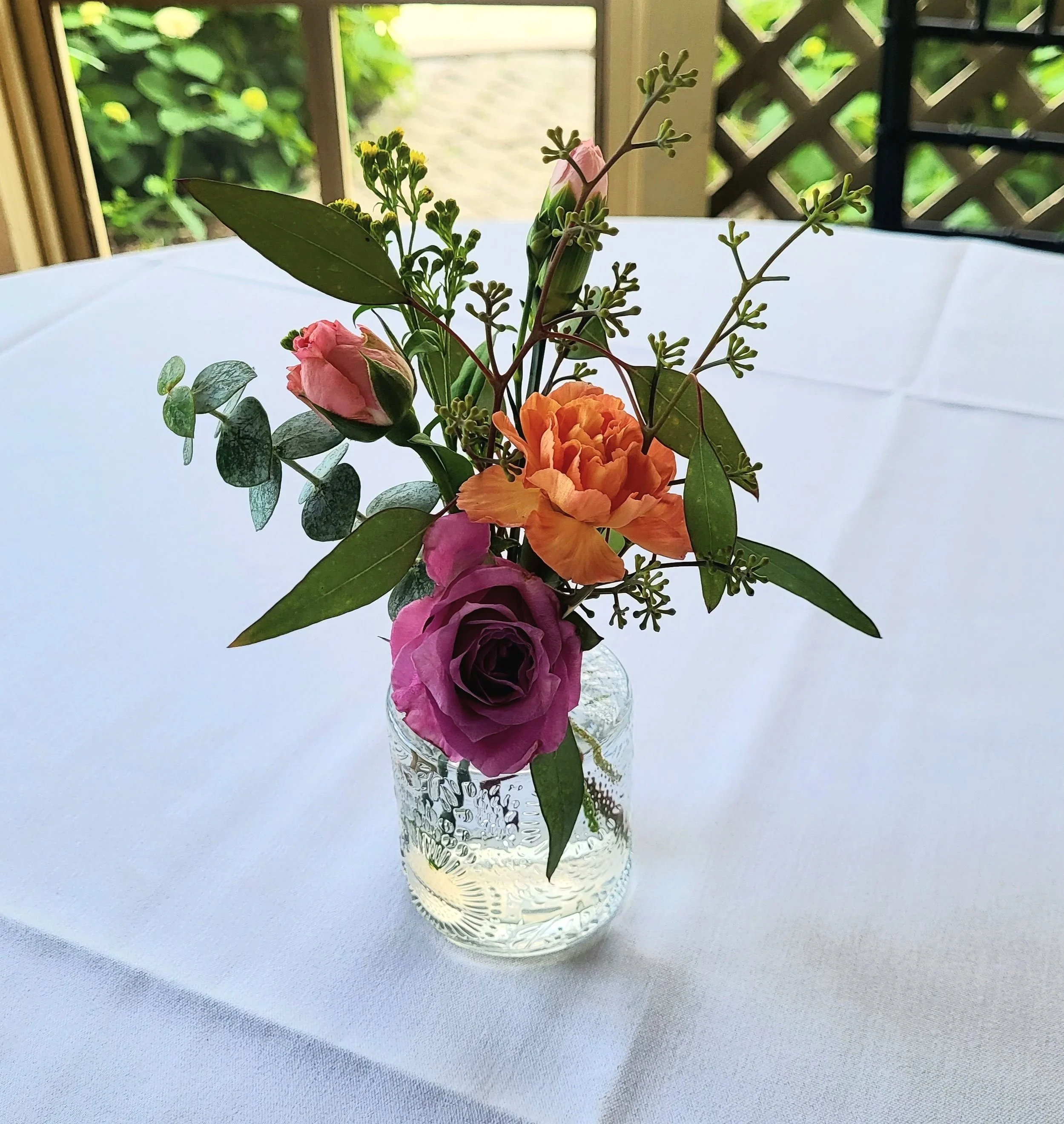 A small glass vase with a textured pattern holds a colorful flower arrangement with pink, orange, and purple flowers and green leaves, placed on a white tablecloth with a window and greenery in the background.