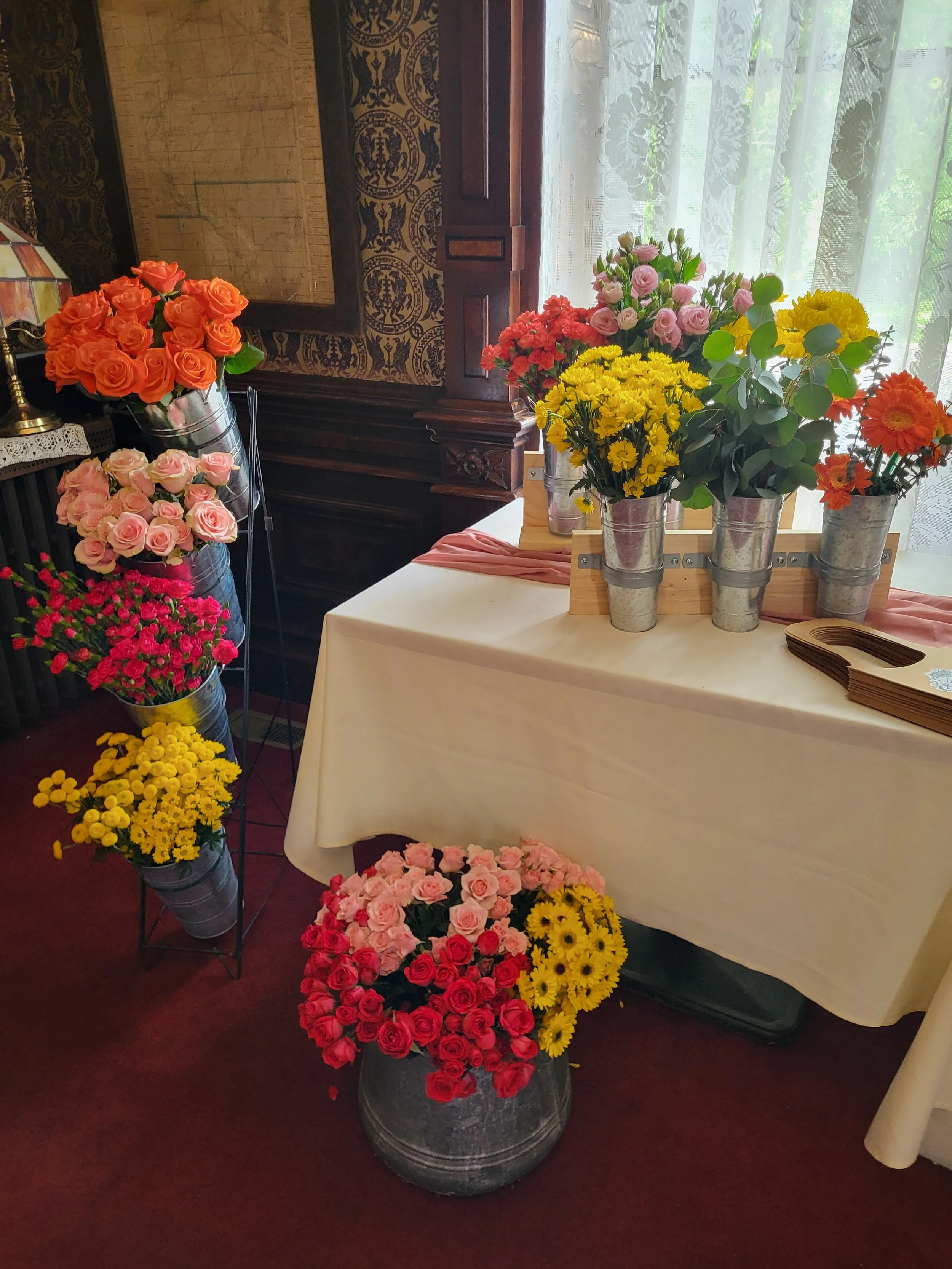 Various colorful flowers in metal buckets displayed on a table and a stand, including pink, yellow, orange, and red roses, daisies, and other blooms, arranged indoors near a window with lace curtains.