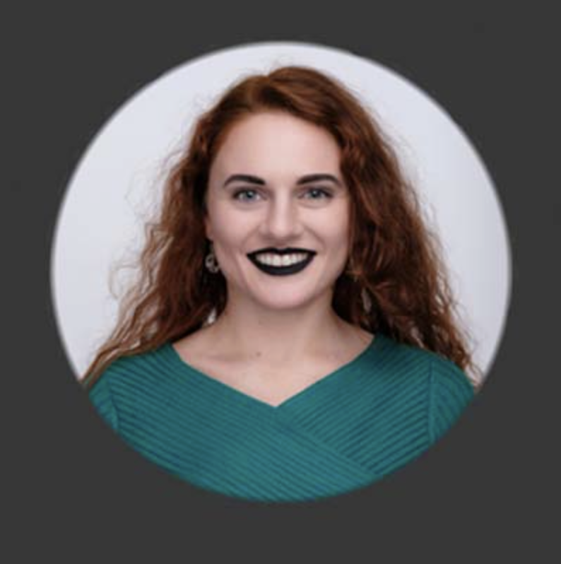 A woman with long red curly hair, black lipstick, and a teal top, smiling against a circular gray background.