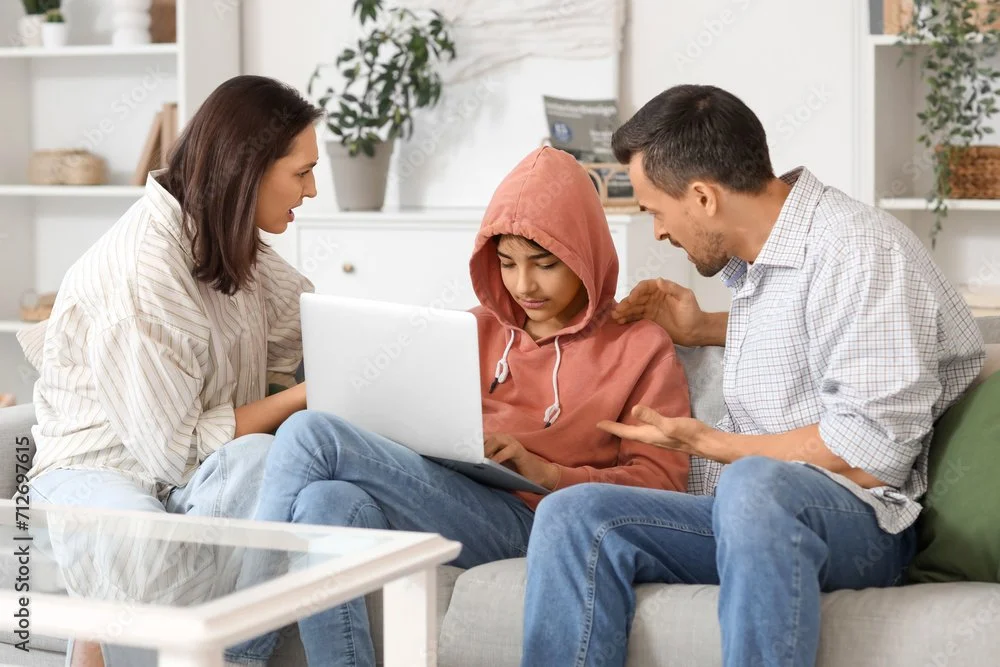A worried young person in a red hoodie sitting on a couch with two adults, one woman and one man, looking at a laptop and a tablet, in a cozy living room with books and plants.