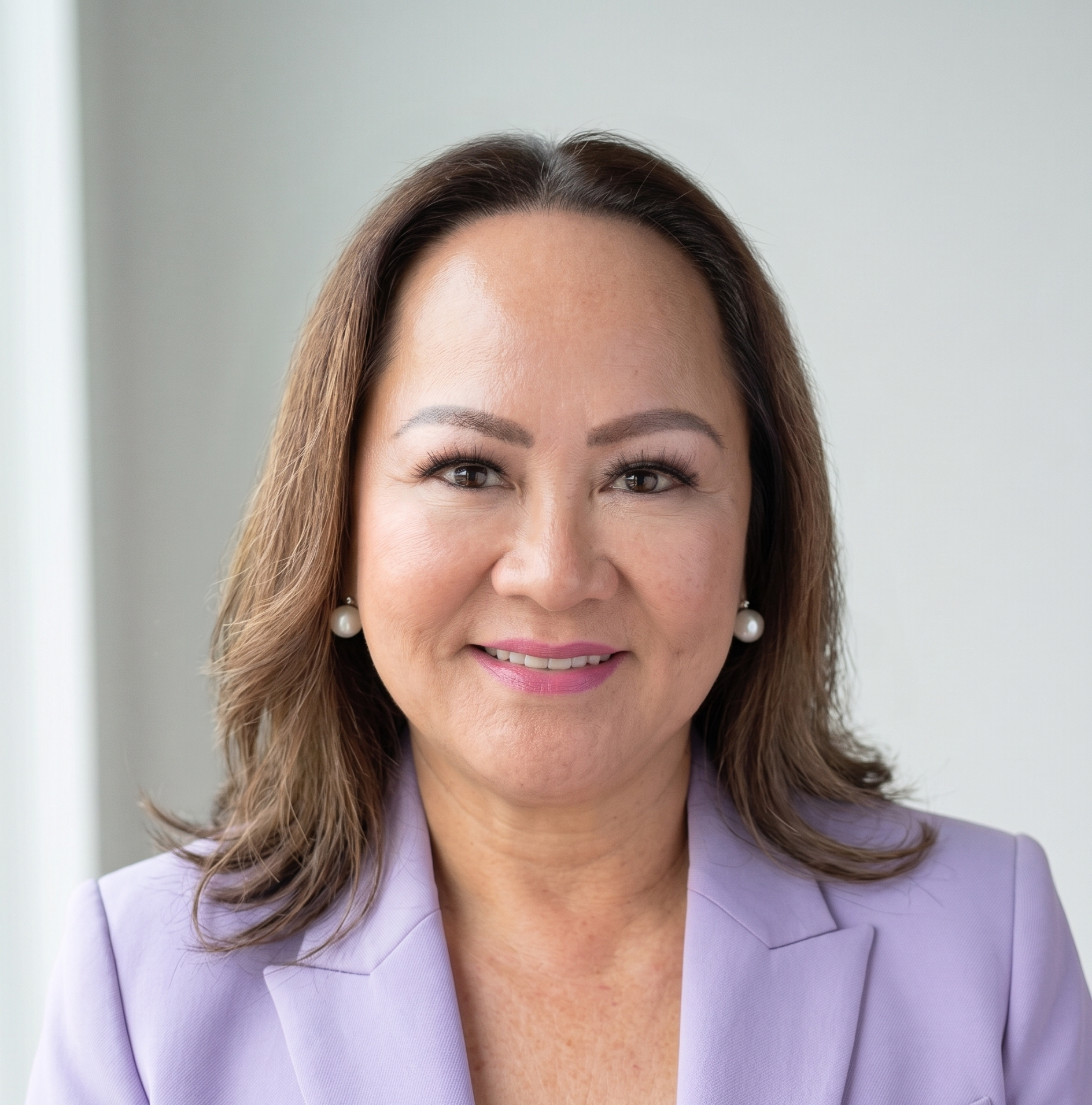 Close-up of a smiling woman with brown hair, wearing pearl earrings and a light purple blazer, in front of a light gray background.