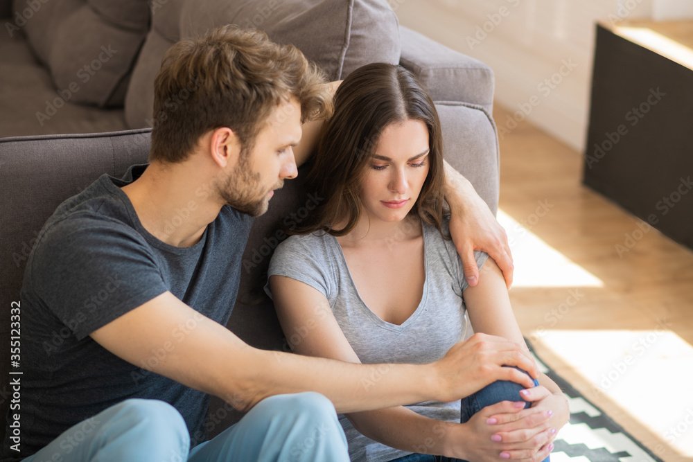 A man gently consoles a woman sitting on a couch, holding her hand, in a cozy living room.