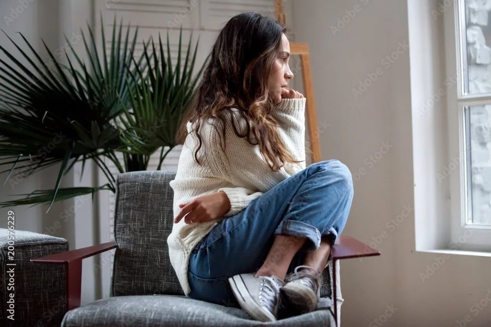 A young woman with long curly hair is sitting on a chair by a window, looking thoughtfully outside. She's dressed in a cream-colored sweater, jeans, and sneakers. There are large green plants and a framed picture in the background in a bright, cozy room.