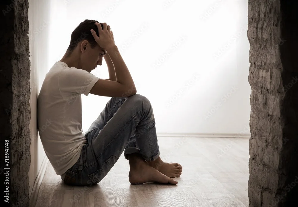 A young man sitting on the floor in a corner with a distressed expression, holding his head in his hands during the daytime.
