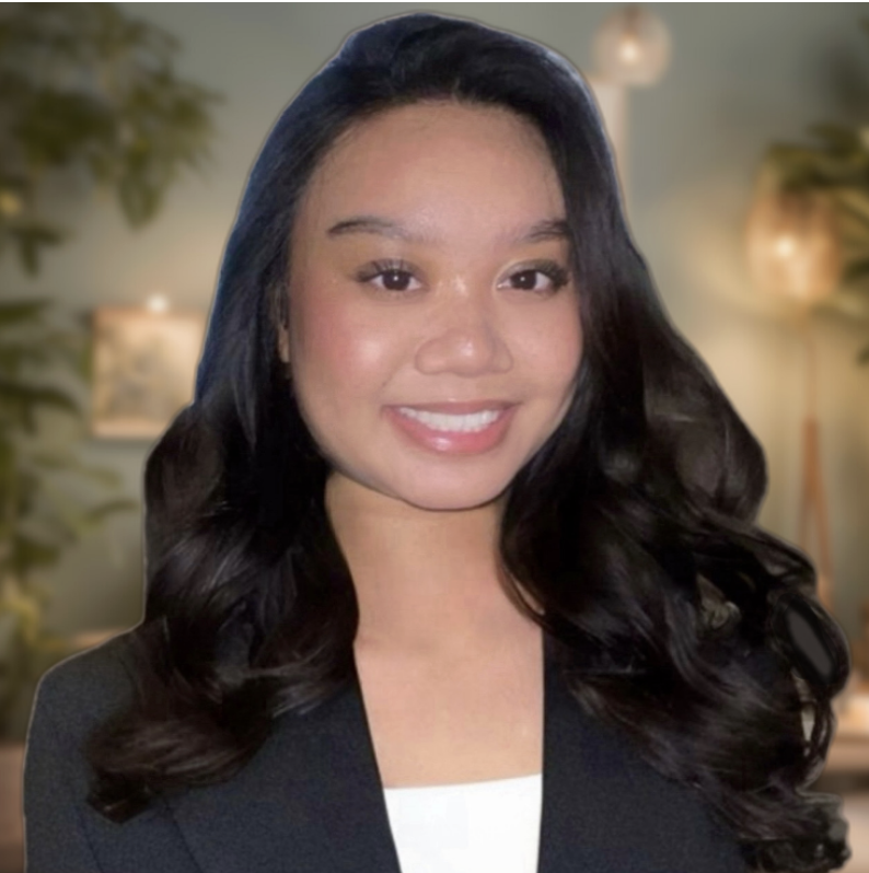 A woman with long dark wavy hair, wearing a black blazer and white top, smiling in a modern, softly lit room with blurred plants and lamps in the background.