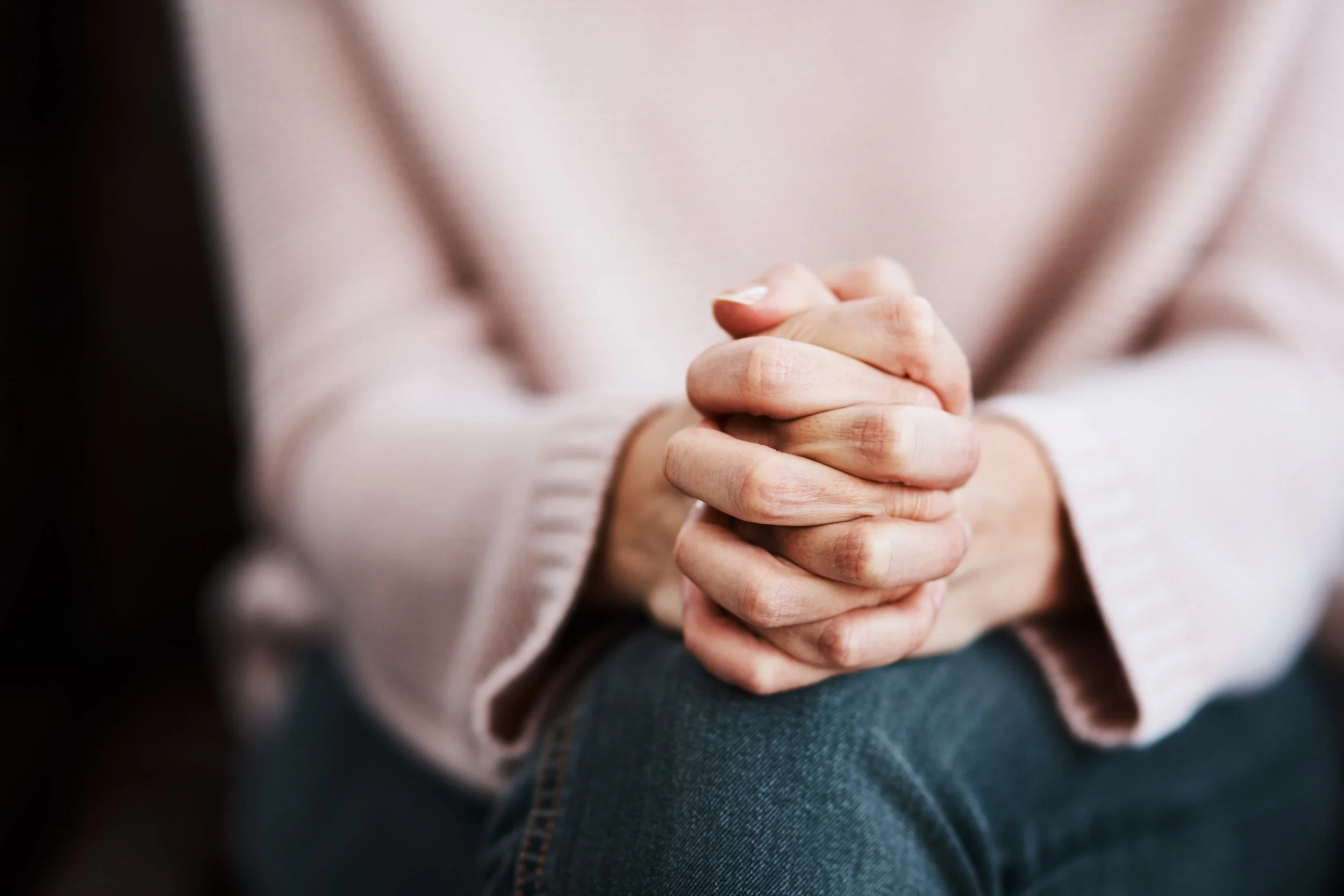 Person with folded hands resting on lap, wearing a light pink sweater and denim jeans.