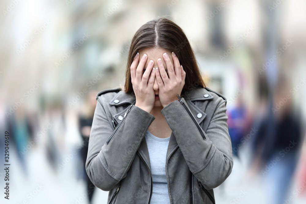Woman holding her face with both hands, standing in a busy indoor setting with blurred people in the background.