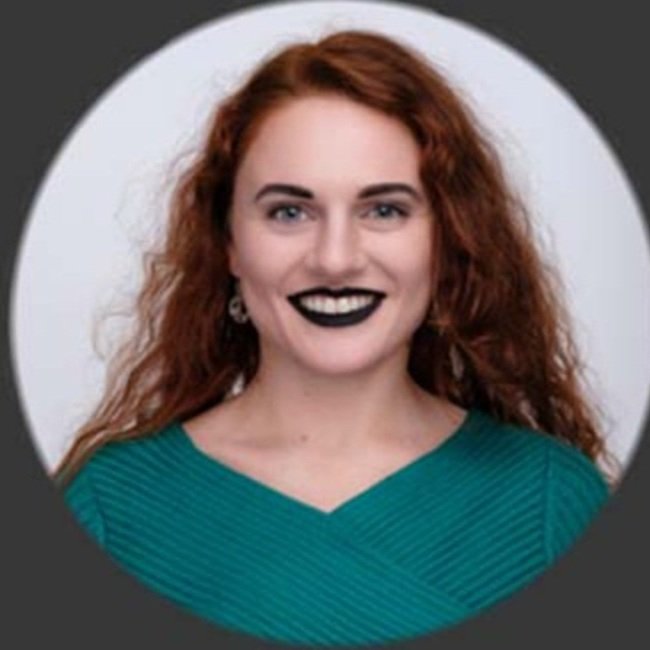 A woman with long, curly red hair, wearing a teal top and black lipstick, smiling at the camera against a white circular background.