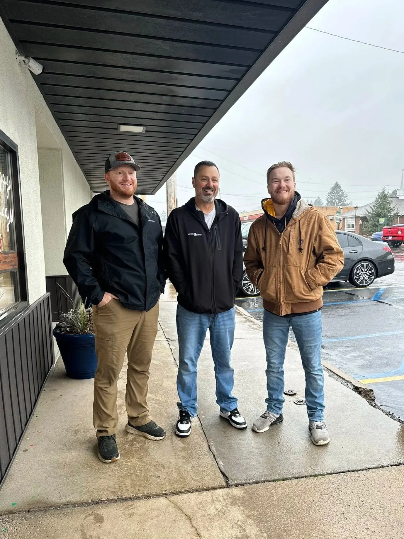 Three men standing outside a building on a wet sidewalk on a cloudy day, smiling at the camera, dressed in casual jackets and jeans.