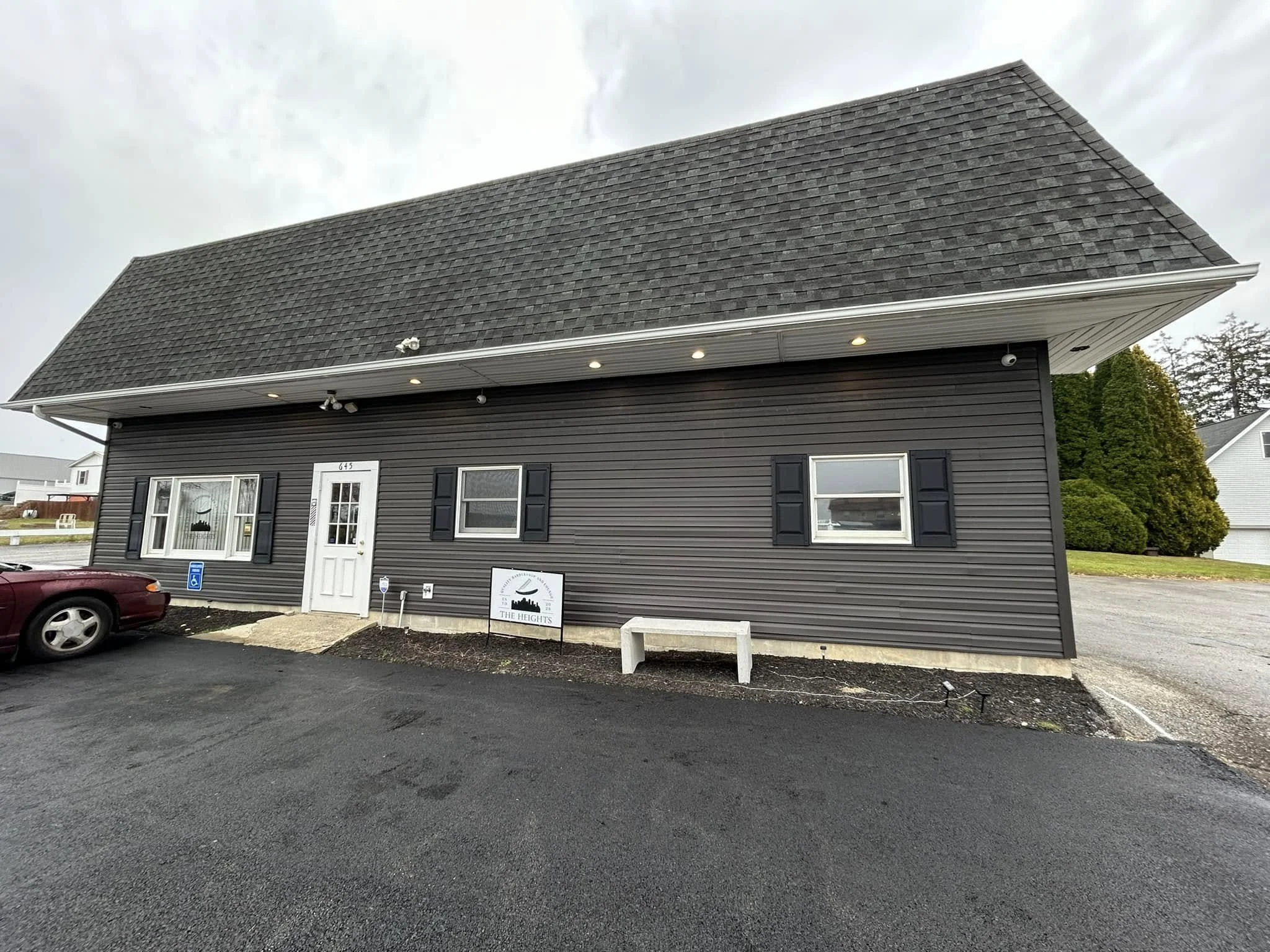 Front view of a modern, dark gray barbershop with black shutters, a white door, and two windows, with a small parking area and a few trees in the background.