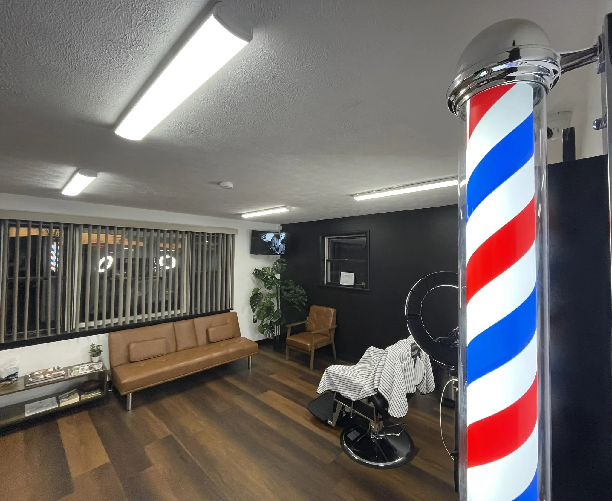 Barber's waiting area with a vintage barber pole, a leather chair covered with a cloth, a small table with magazines, a leather sofa, an armchair, a potted plant, and a TV on the wall.