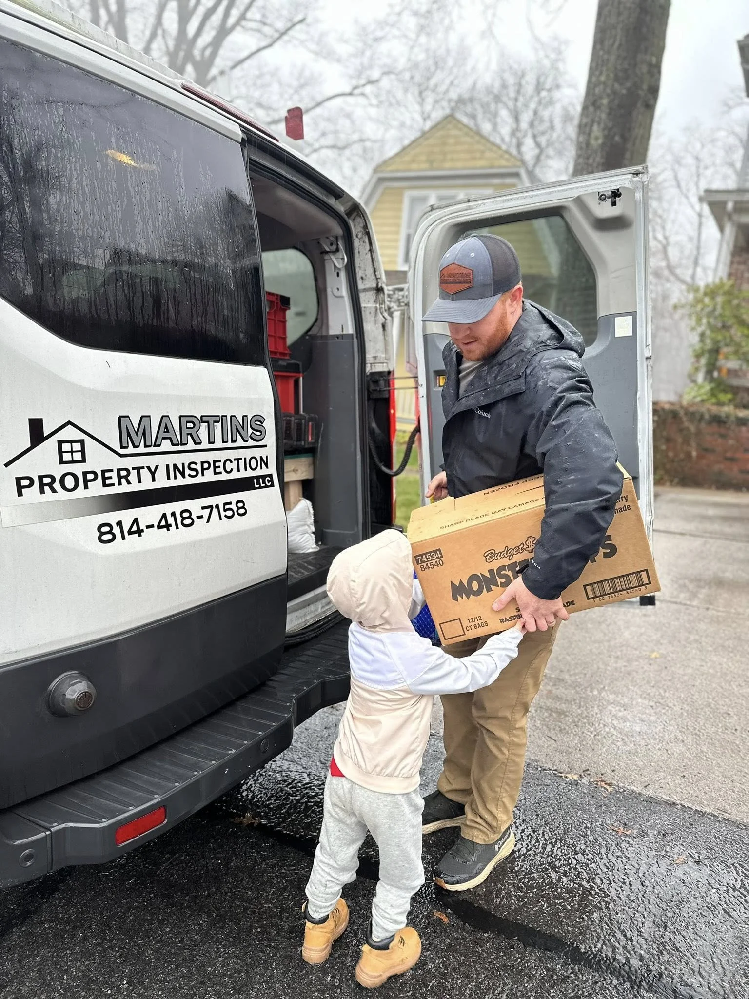 A man delivering a box of snacks to a young child standing next to a delivery van that reads 'Martins Property Inspection LLC'.