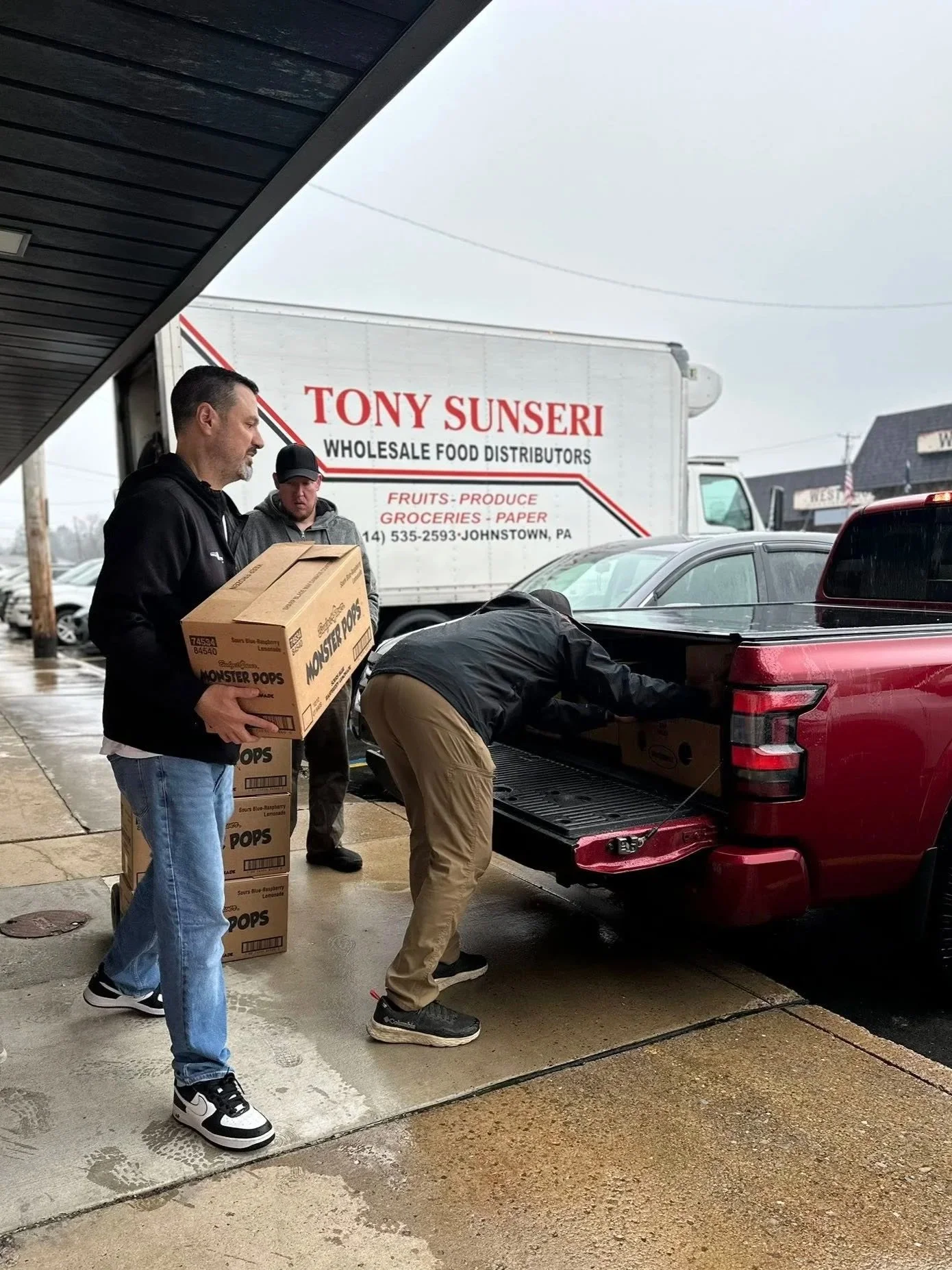 Three men unloading boxes of Monster Pops ice cream from a red pickup truck outside a wholesale food distribution store on a rainy day.