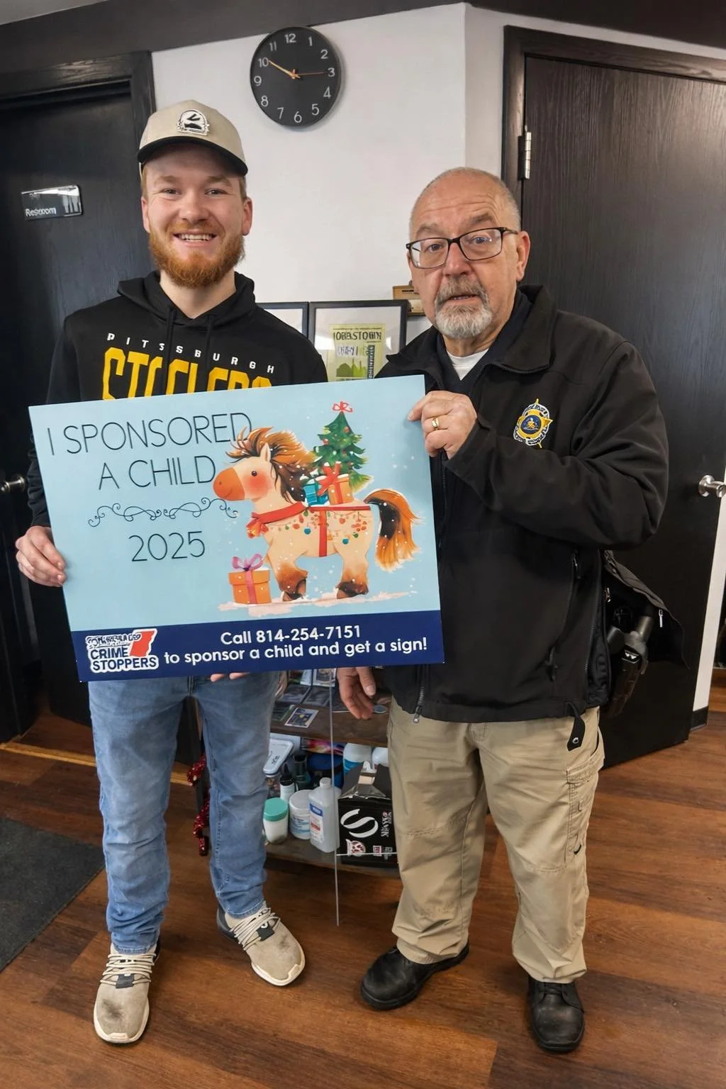 Two men standing indoors, holding a large Christmas-themed sign that reads 'I Sponsored a Child 2025' with a cartoon horse and Christmas decorations. The older man is wearing a black jacket with a police badge, glasses, and beige pants. The younger man is wearing a beige cap, black hoodie with Pittsburgh Steelers logo, and jeans. A clock on the wall shows 2:55.