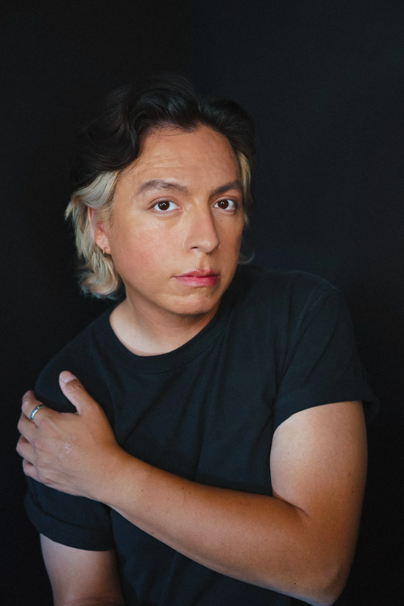 A portrait of Willi White with short black and blonde hair, wearing a black shirt, looking at the camera, against a black background, with arms crossed.