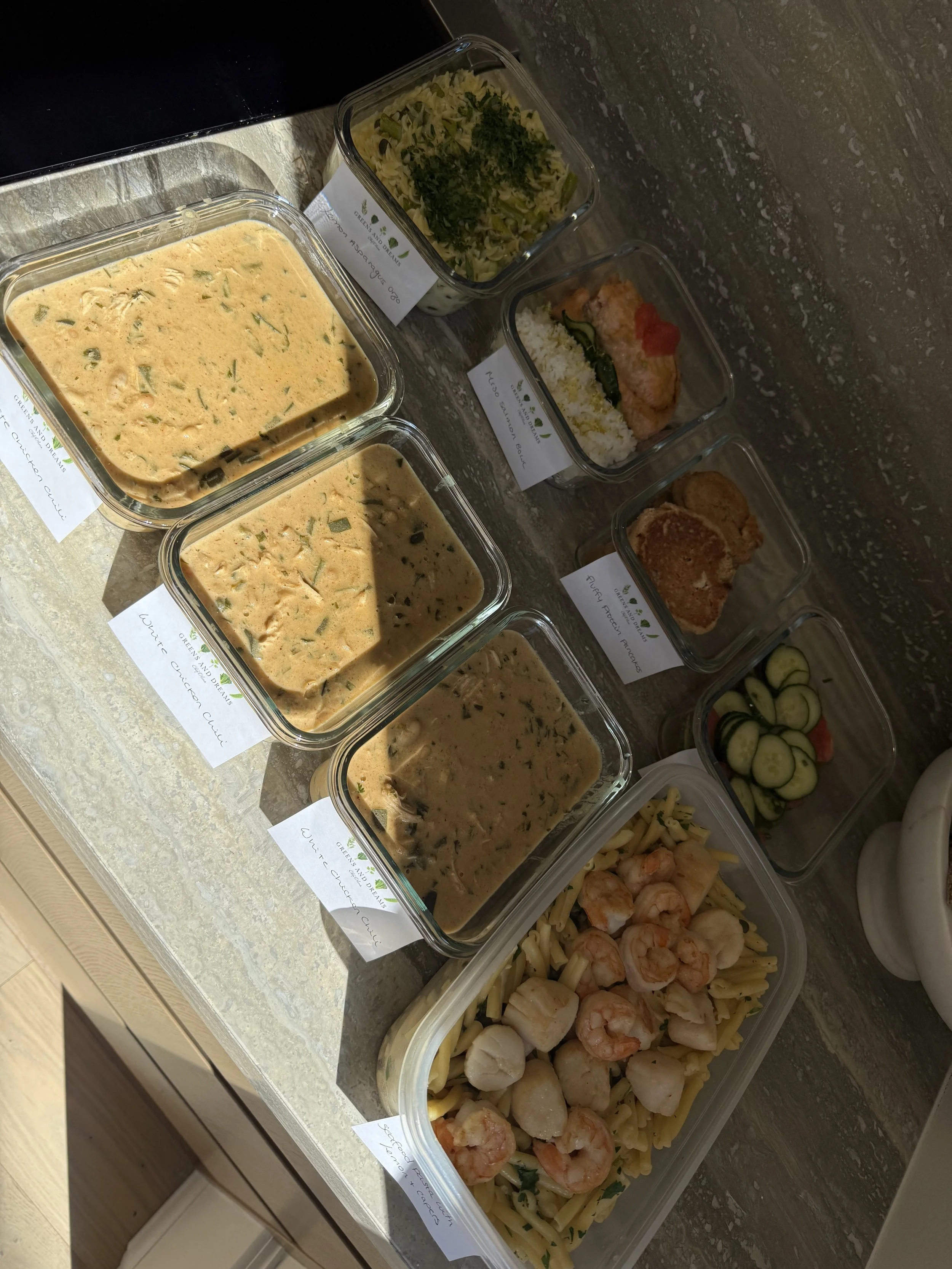 Several glass containers with different types of pasta and sauces, including a creamy sauce with green herbs, and other containers with cooked pasta, shrimp, scallops, cucumber slices, and cooked chicken, indicating a prepared meal or ingredients for