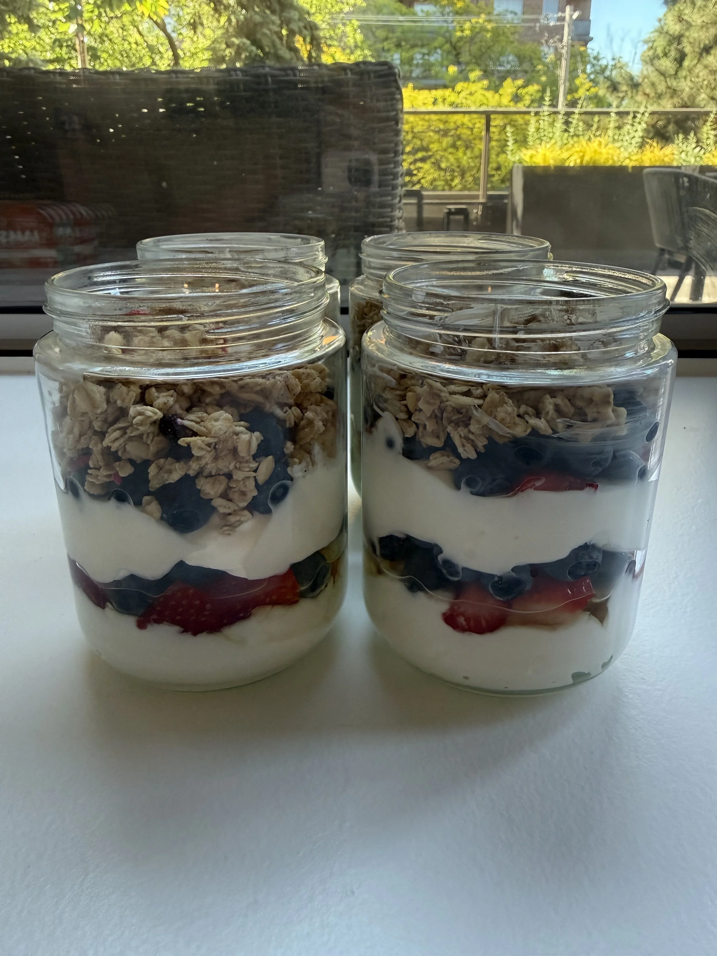 Four glass jars filled with layers of yogurt, granola, blueberries, and sliced strawberries on a white surface with a window showing trees and outdoor seating in the background.