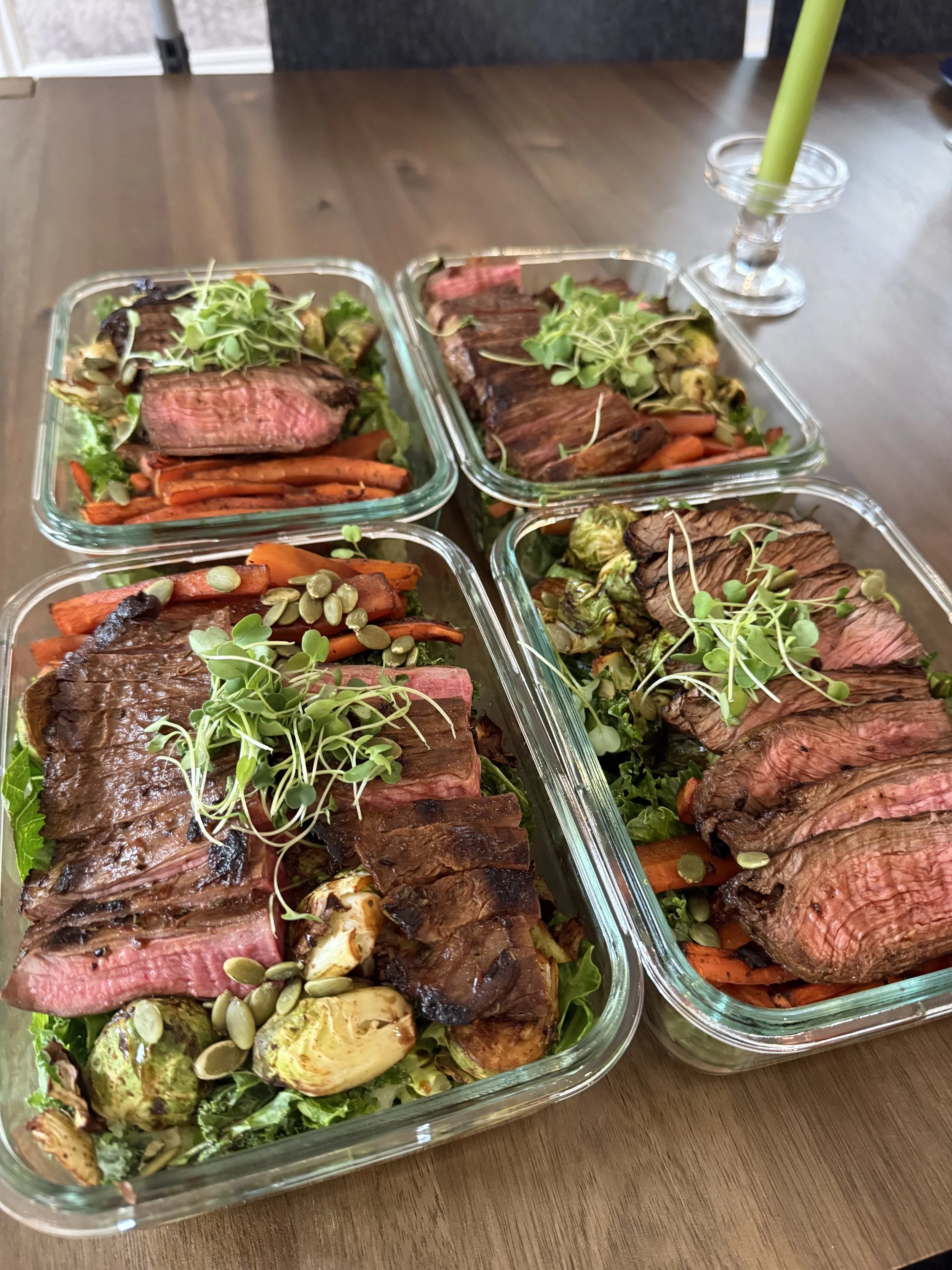 Glass meal prep containers filled with sliced grilled steak, roasted carrots, roasted Brussels sprouts, and garnished with microgreens on a wooden table.