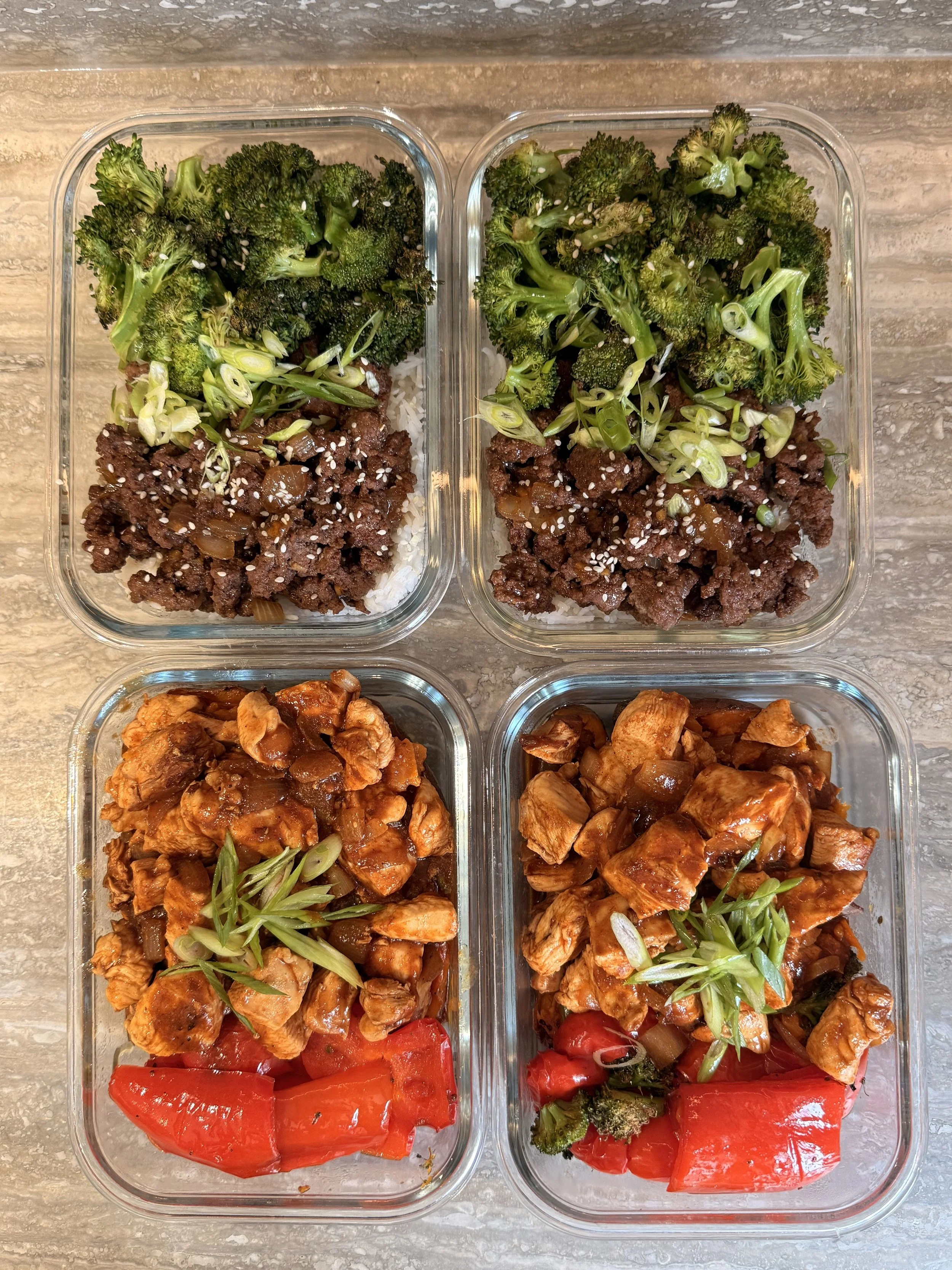 Two glass meal prep containers filled with rice, raw broccoli, ground beef, chicken, and red bell peppers, with slices of green onion on top of the beef and chicken, set on a gray countertop.