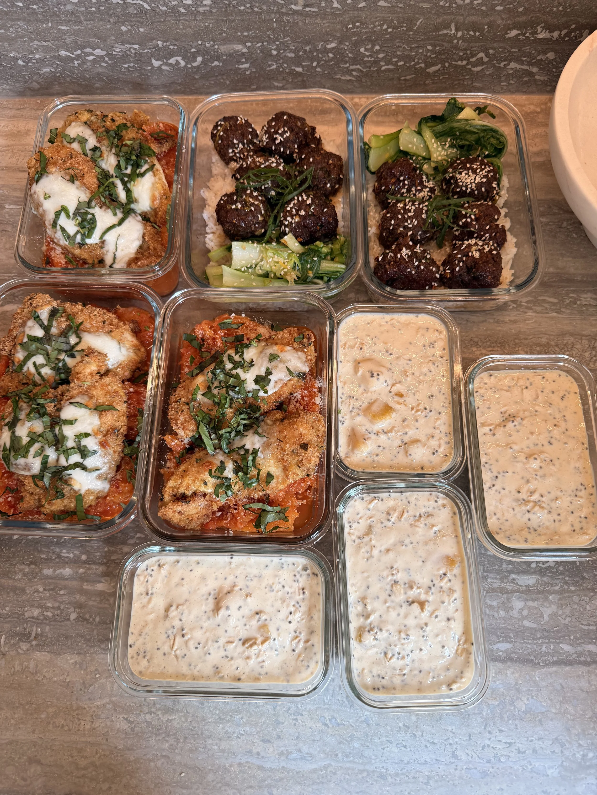 Assorted meal prep containers with baked chicken topped with herbs, meatballs with cucumber, and creamy sauces on a gray marble countertop.