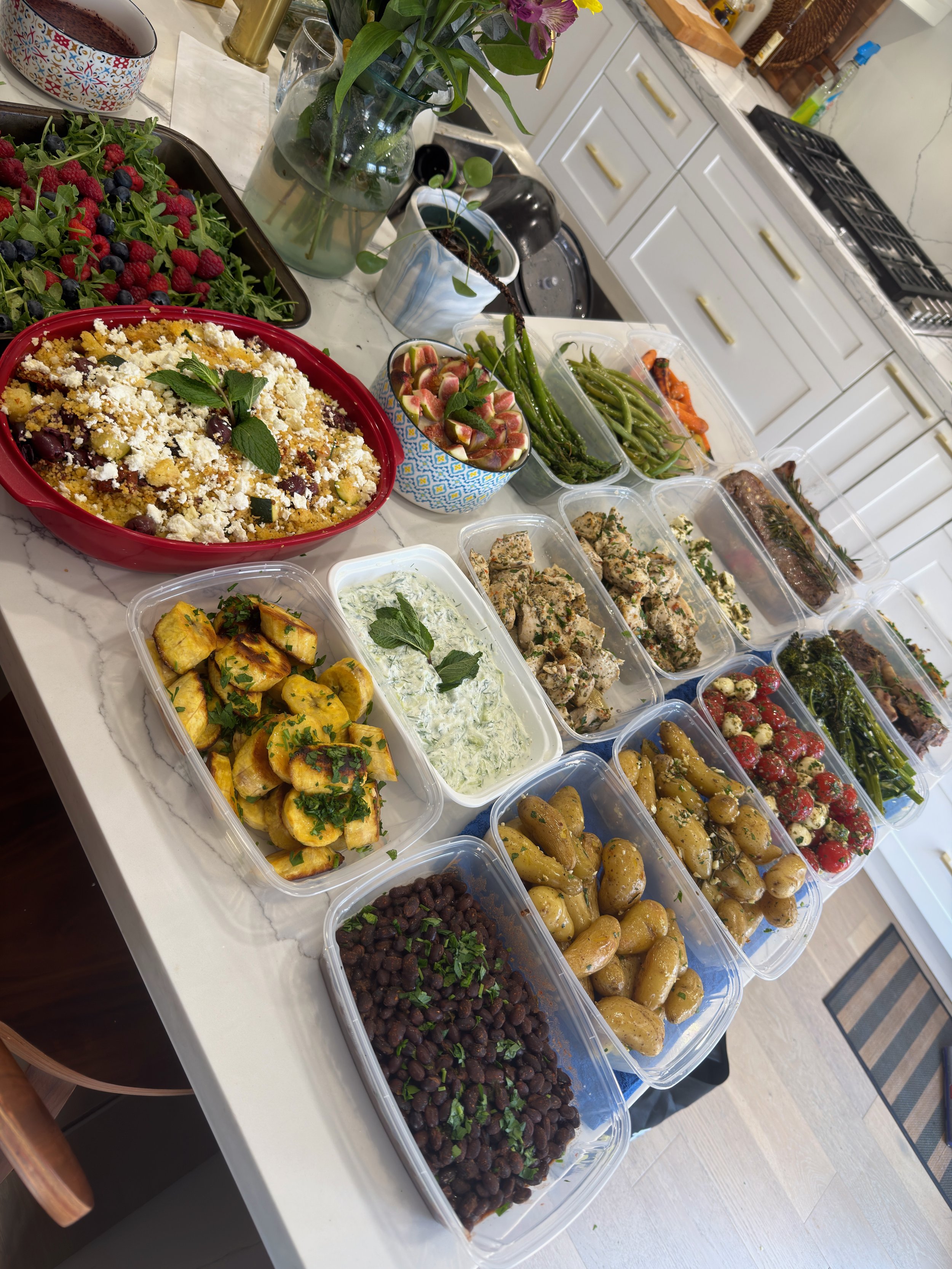 A kitchen counter with various prepared dishes in plastic containers, including roasted potatoes, salads, grilled vegetables, and a fruit salad, with a large fruit and vegetable salad, a flower vase, and other items in the background.