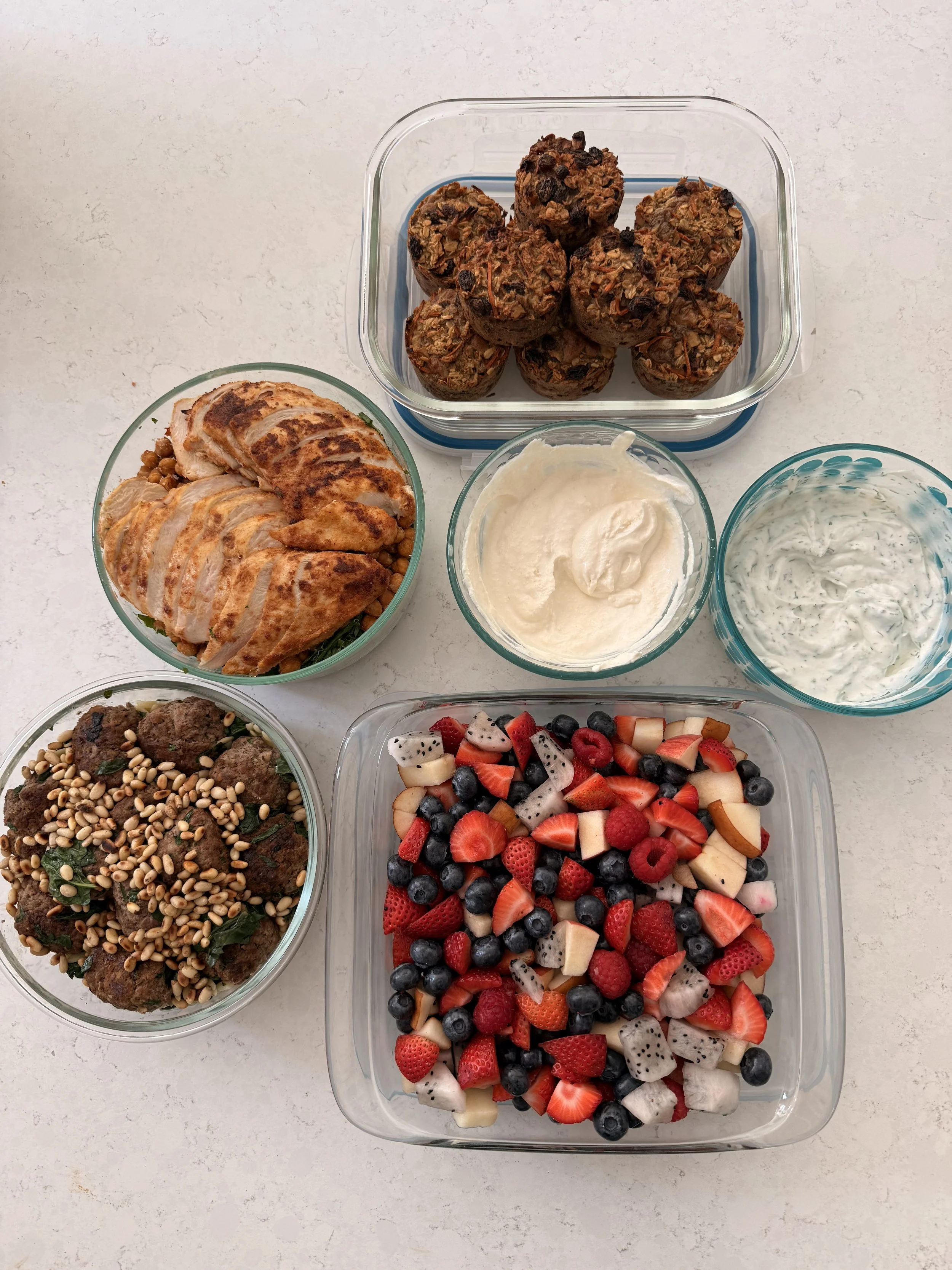 Assorted desserts and fruit bowls on a light-colored countertop, including oatmeal muffins with chocolate chips, grilled chicken slices, creamy dips, and a mixed fruit salad with strawberries, blueberries, raspberries, and chopped exotic fruits.