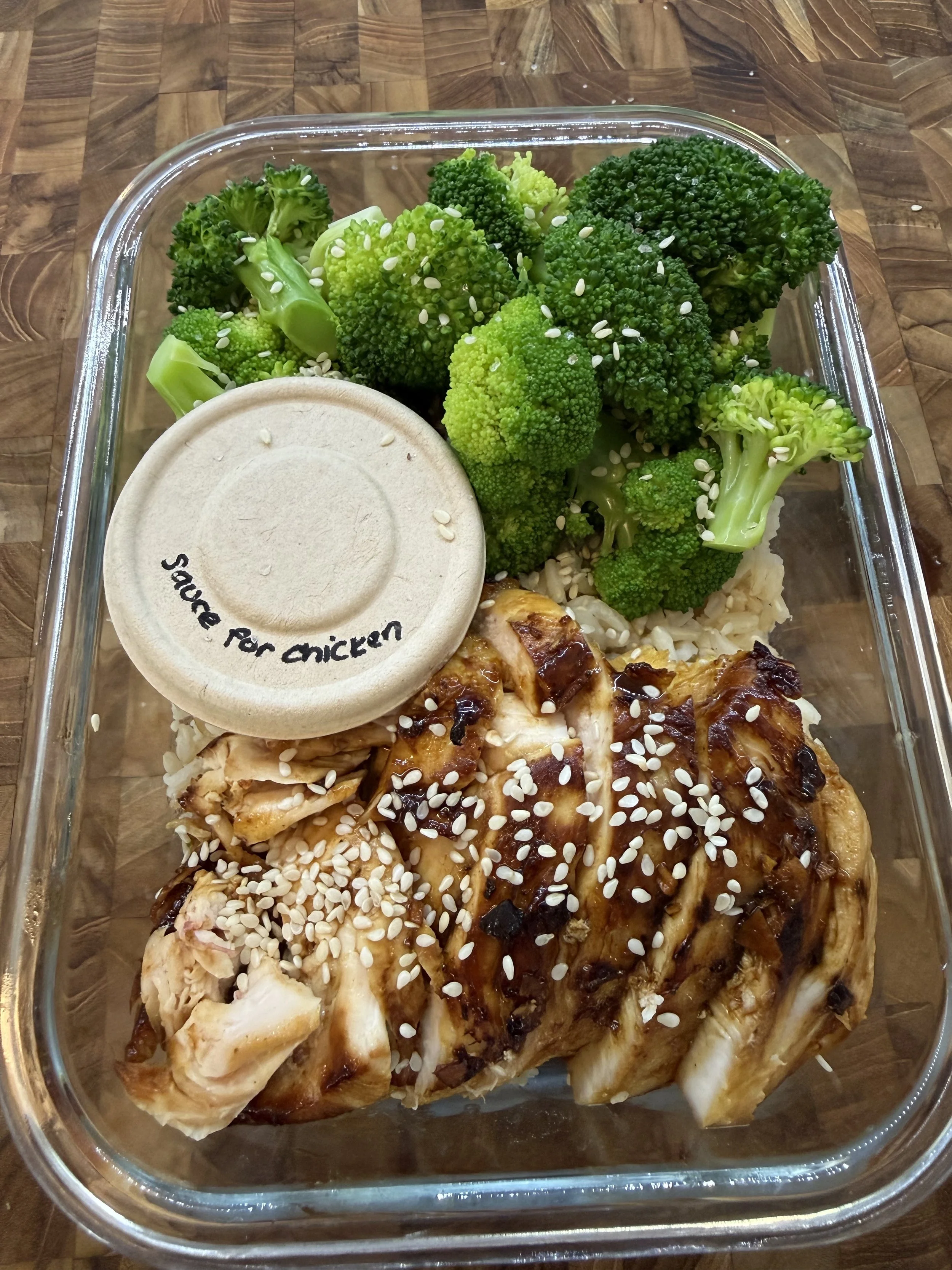Cooked chicken with barbecue sauce and sesame seeds, broccoli florets, and rice in a glass container.