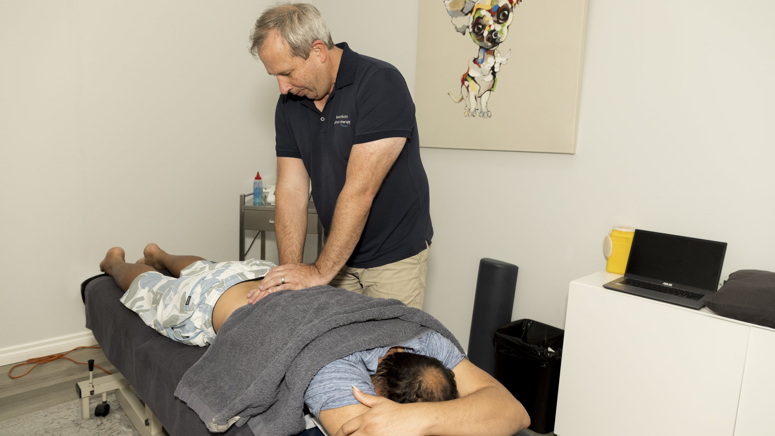 A chiropractor performing an adjustment on a patient lying face down on a treatment table in a clinic room, with a colorful dog painting on the wall behind them.