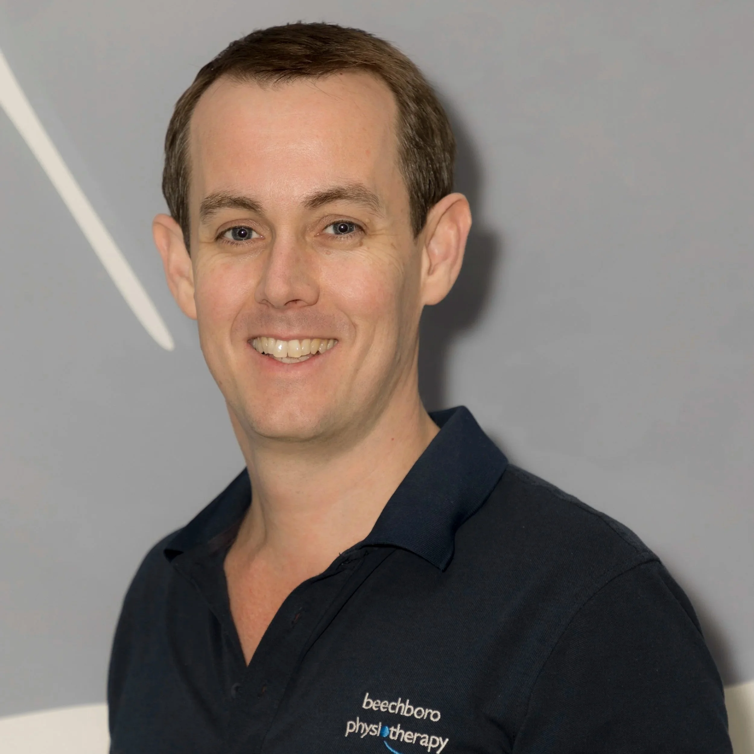 Kevin Forrest in front of a gray background, wearing a navy blue polo shirt with the logo 'beechboro physiotherapy'.