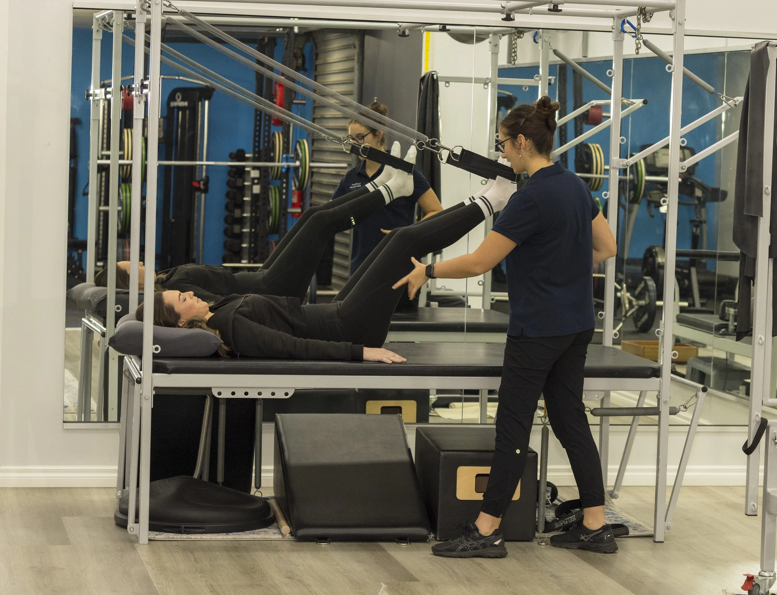 A woman lying on a therapy table doing leg exercises with straps, while a therapist assists her with her stretching session in a gym or physical therapy room.