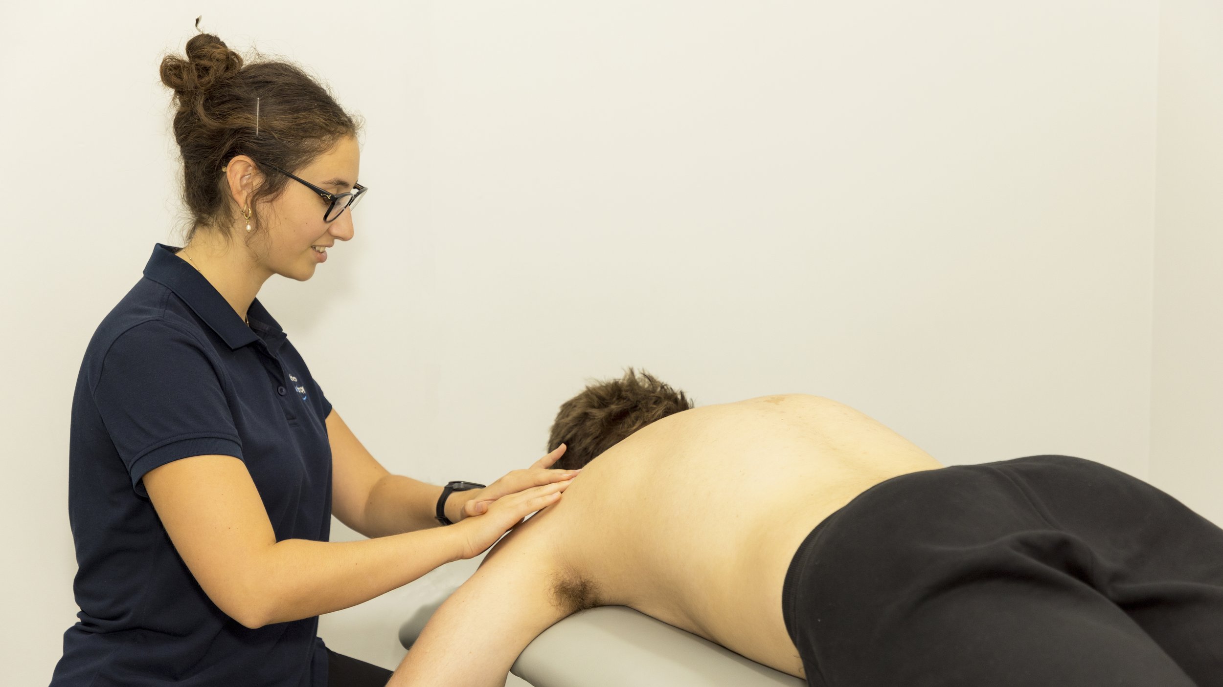 A female therapist in glasses wearing a navy polo shirt providing physical therapy or massage to a male patient lying face down on a treatment table, in a clinical setting with plain white walls.