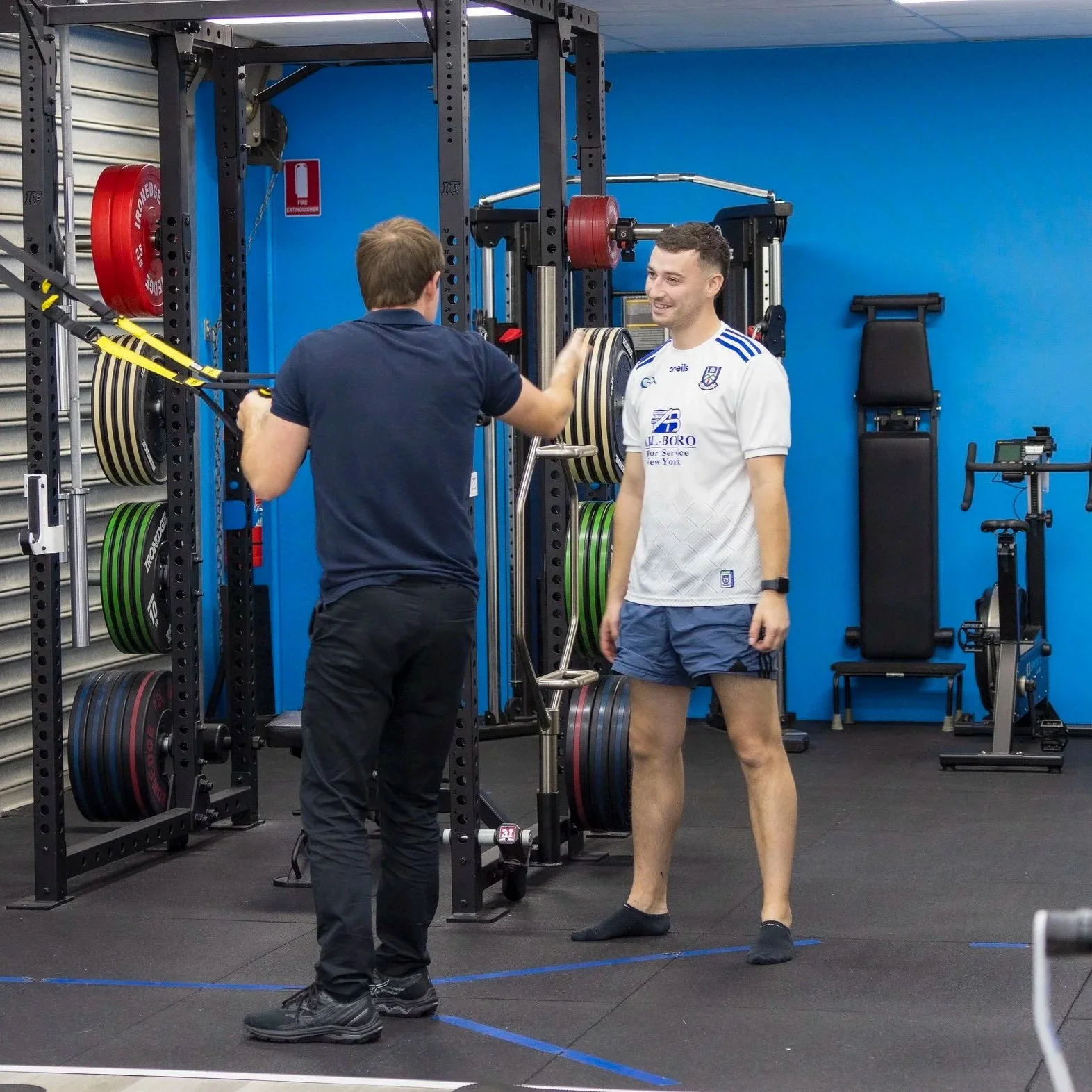 Two men in a gym standing next to a weightlifting rack. One man is wearing a sports jersey and shorts, smiling, while the other man is wearing dark clothing and appears to be instructing or conversing with him. The gym has various weights and exercise equipment.