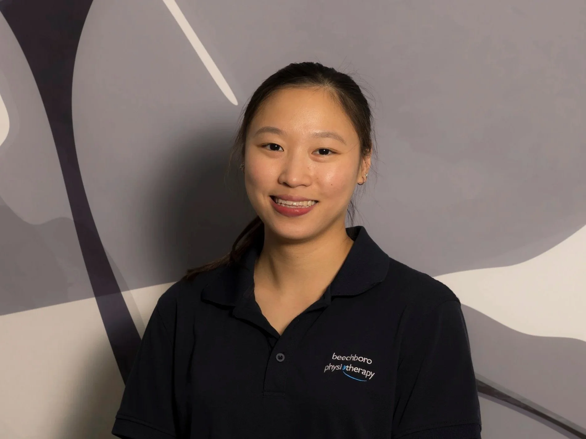 Young woman with dark hair tied back, wearing a black polo shirt with 'beechboro physiotherapy' embroidered on it, smiling in front of a gray and white abstract background.
