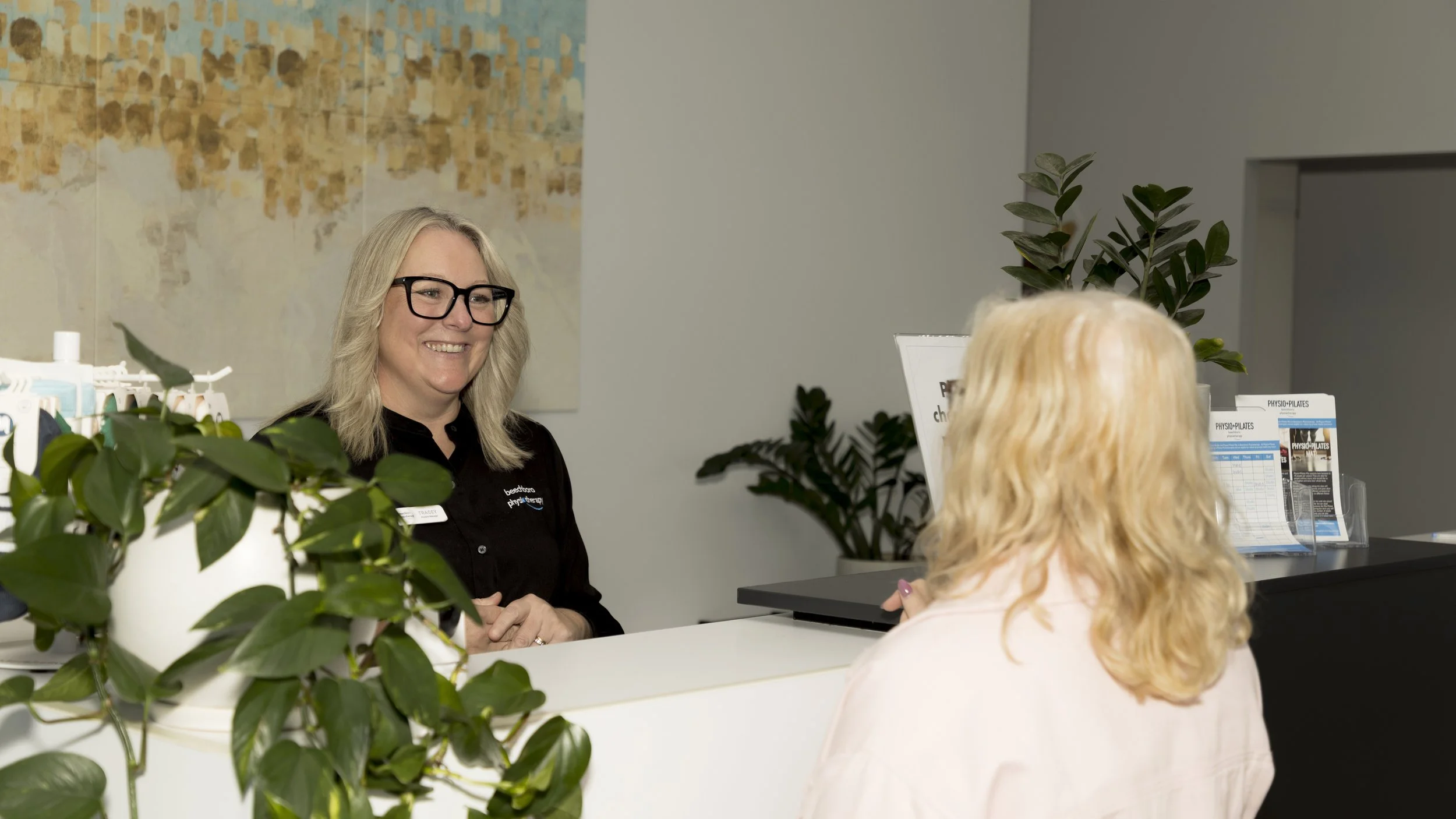 A woman with blonde hair wearing glasses and a black shirt smiling at a woman with blonde hair sitting at a reception desk.