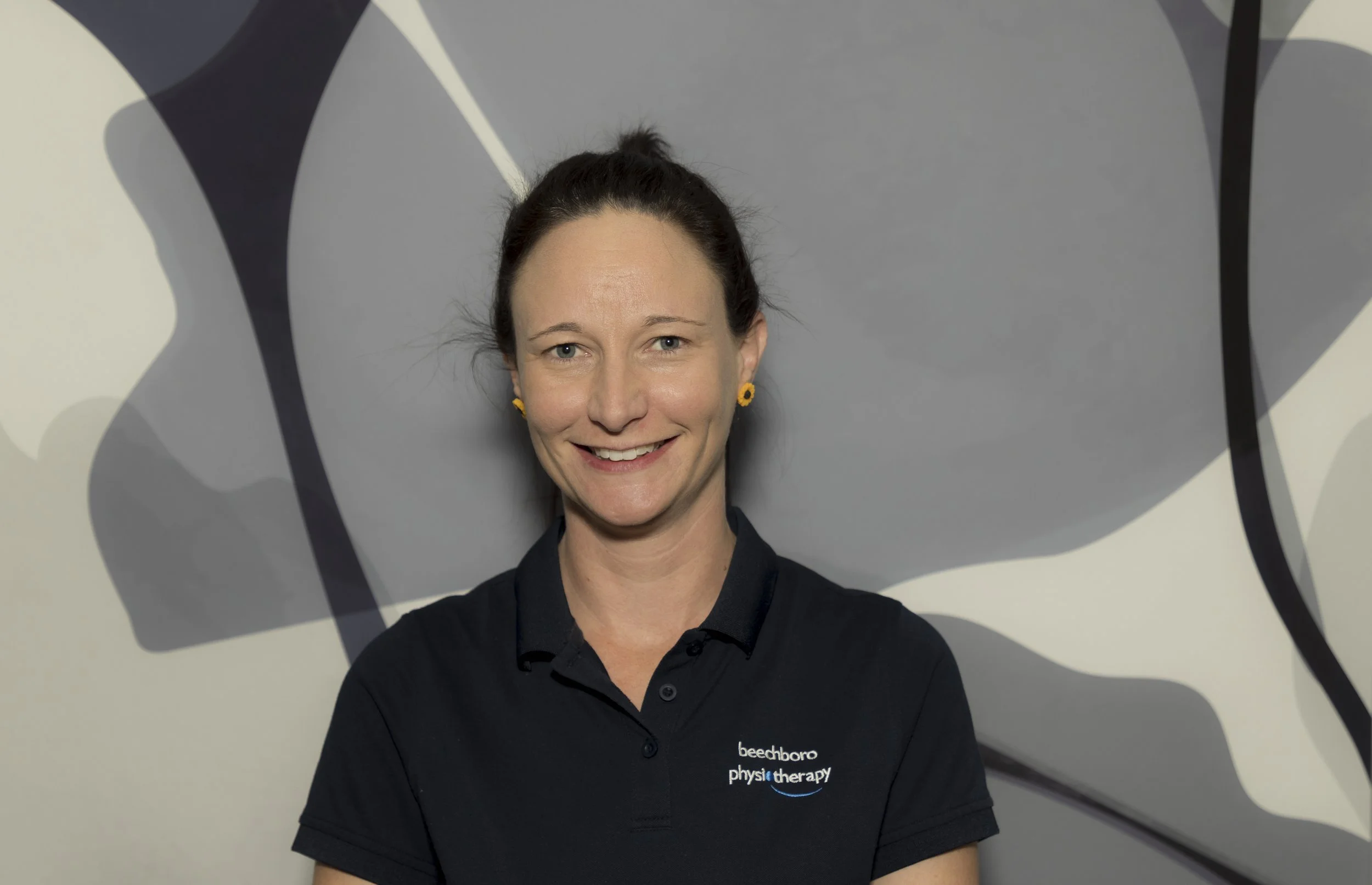 A woman with dark hair tied back, wearing a black polo shirt with a logo, smiling and standing in front of an abstract black and white background.