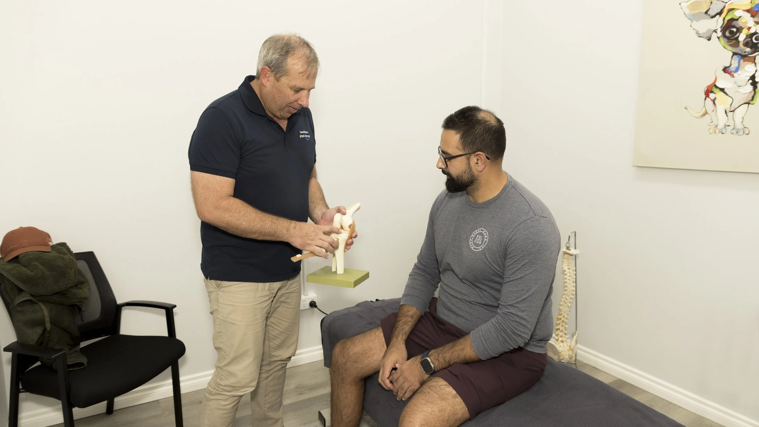A healthcare provider explains a knee joint model to a male patient sitting on an examination table in a medical office.