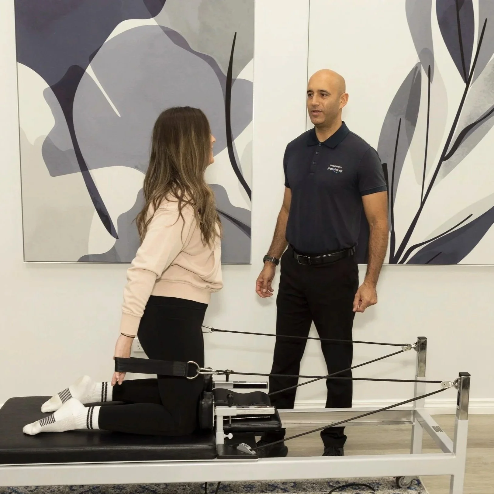 A woman with her knee attached to a scientific device, kneeling on a padded table, having a conversation with a male physical therapist or doctor in a clinical setting.