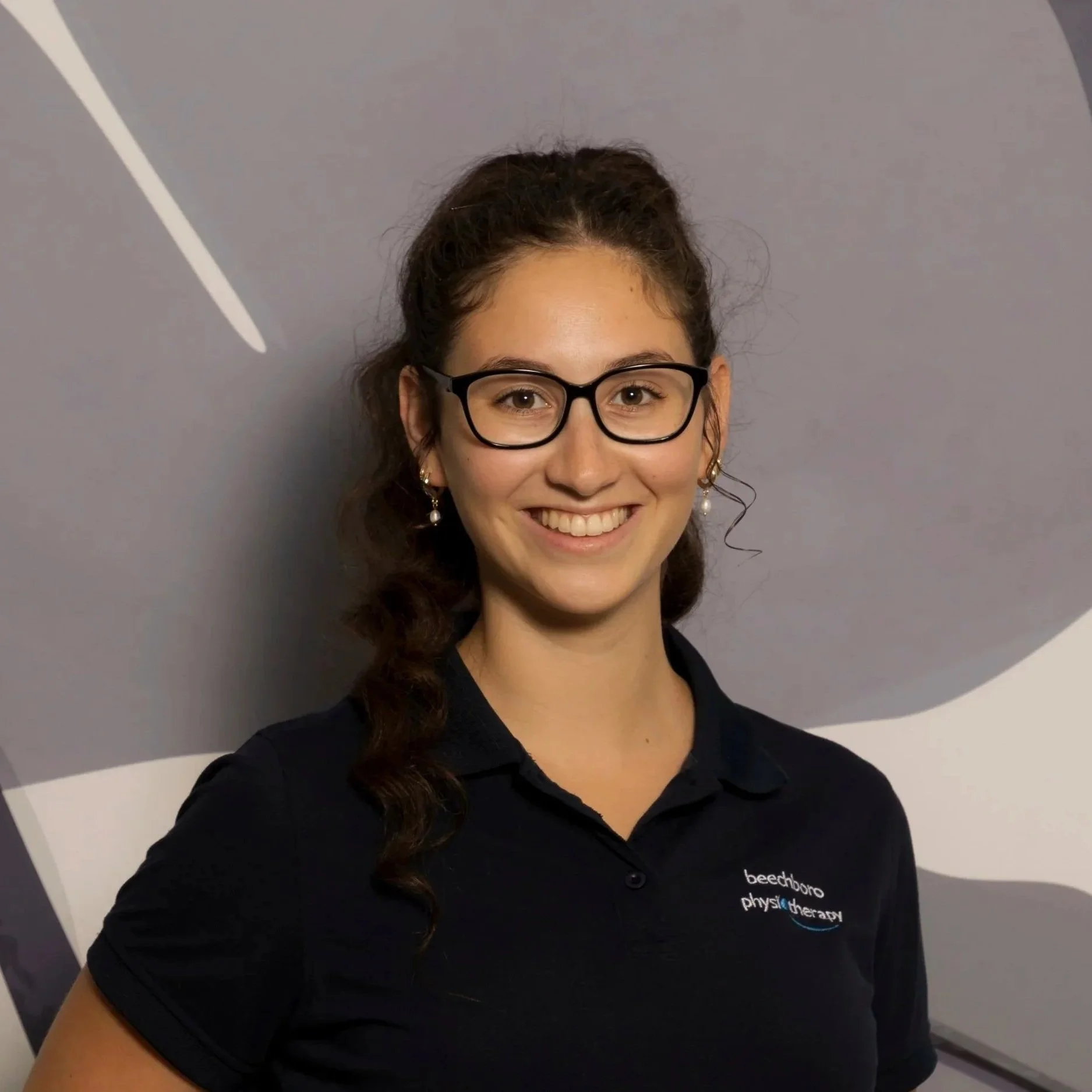 Abi Gardiner against a gray background with abstract white and purple shapes. She is wearing a black polo shirt with a logo for 'beechboro physiotherapy'.