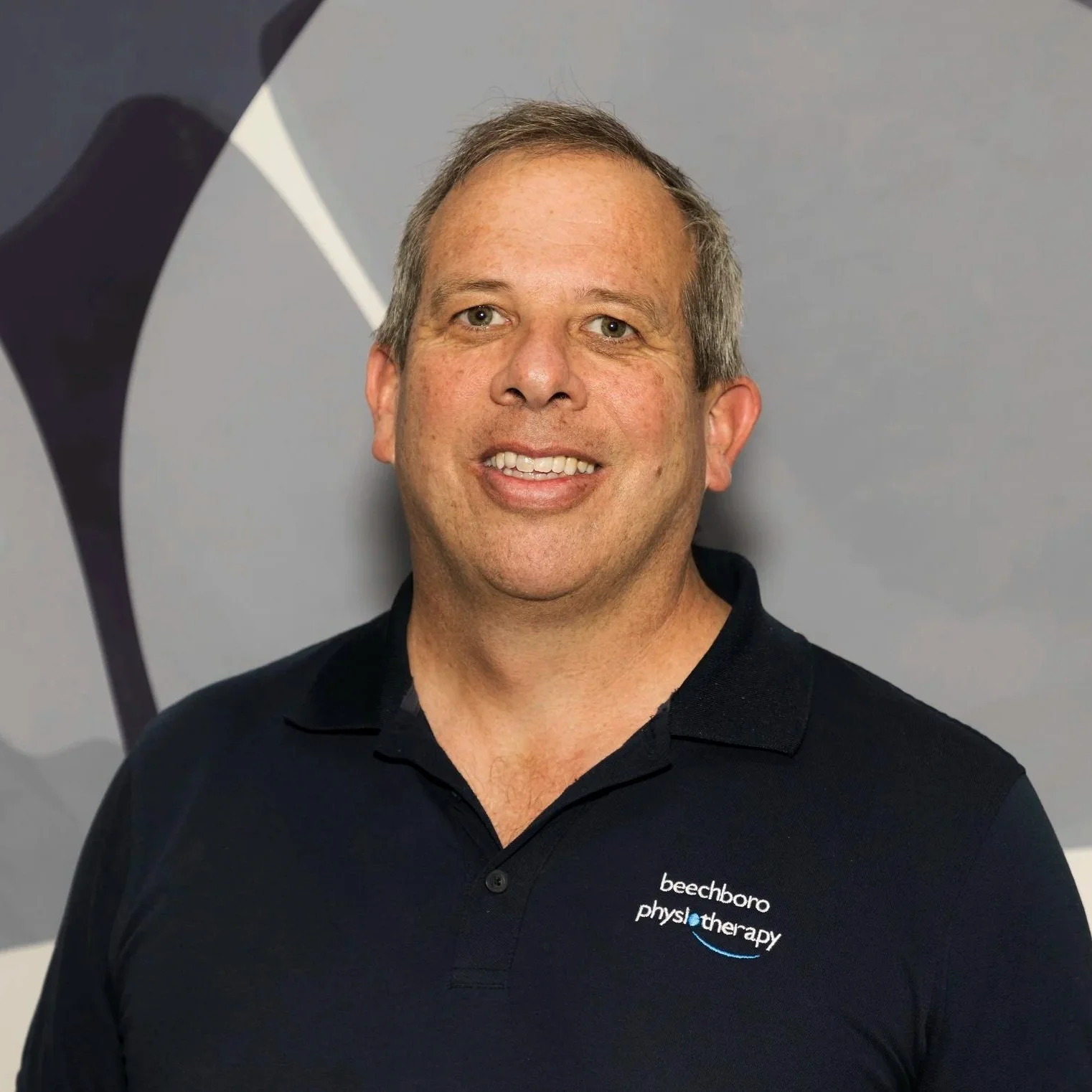 Grant Collins in front of a gray background with a black and gray abstract design. He is wearing a black polo shirt with the words 'beechboro physiotherapy' embroidered on it.