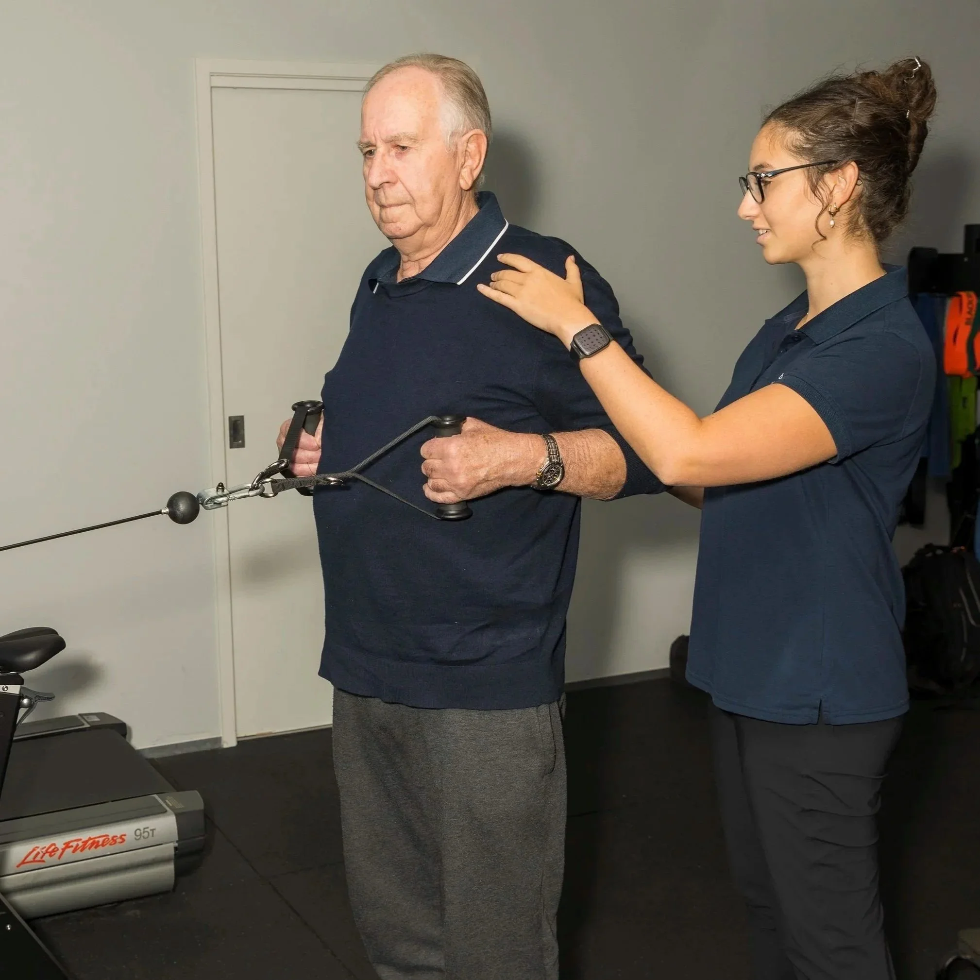 A young woman assisting an elderly man with a resistance training exercise using a cable machine at a gym.