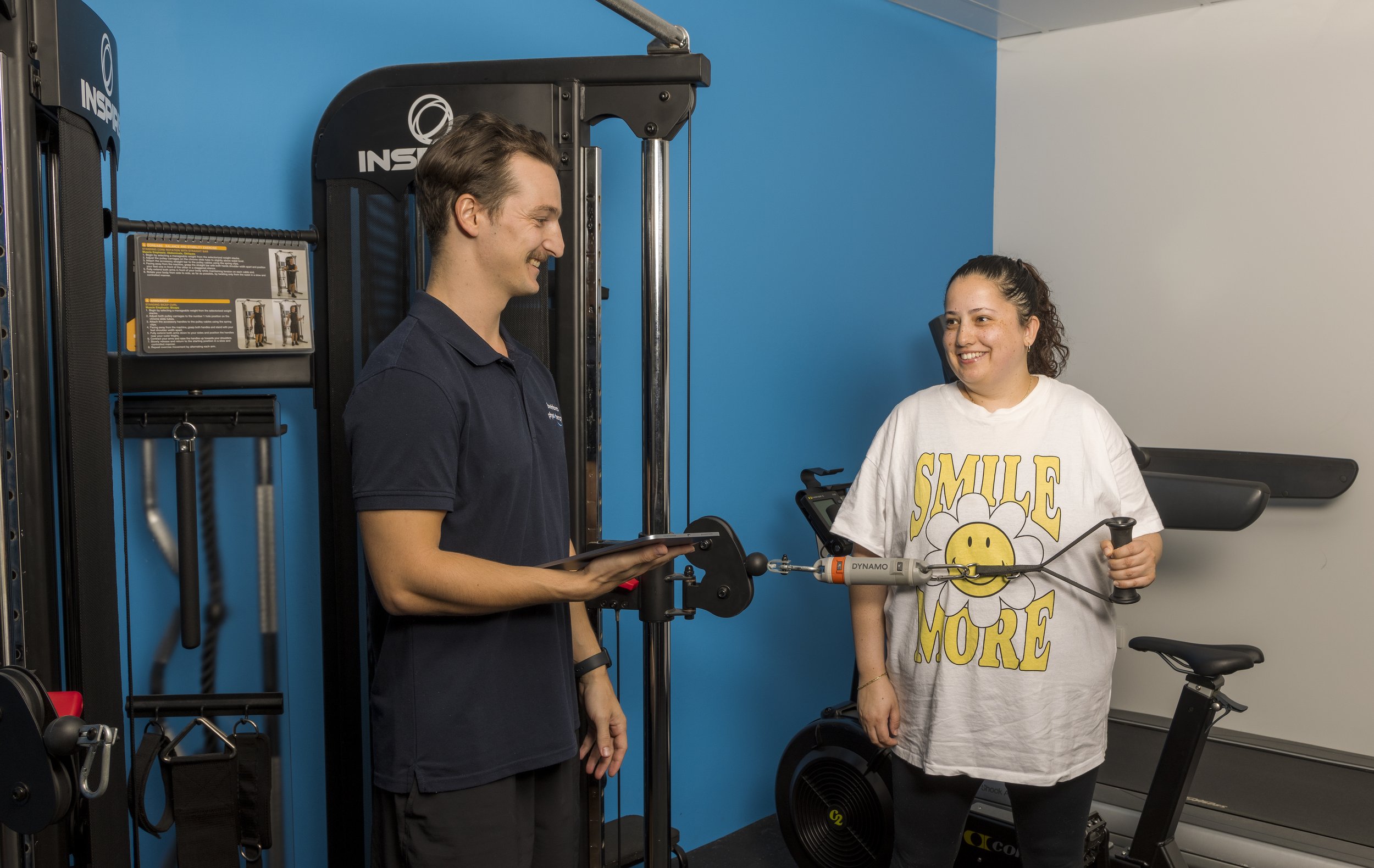A woman is using resistance exercise equipment while smiling at a fitness trainer at a gym. The trainer is holding a tablet and smiling back. The woman is wearing a white t-shirt with the words 'Smile More' and a smiley face graphic, and the gym has blue walls.