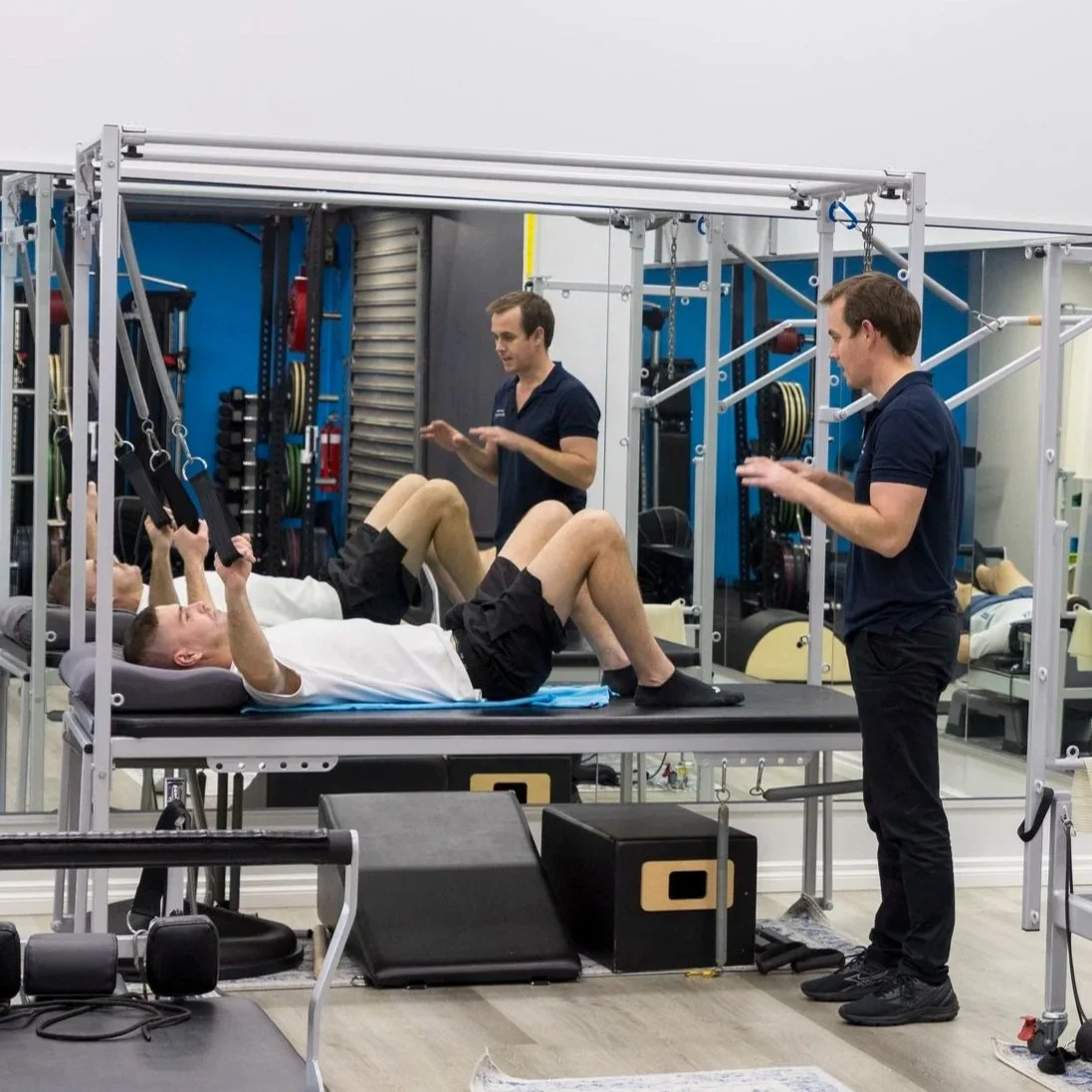 A person lying on a therapy table engaging in physical therapy exercises with resistance bands, while two therapists assist and instruct in a rehabilitation facility with various exercise equipment in the background.