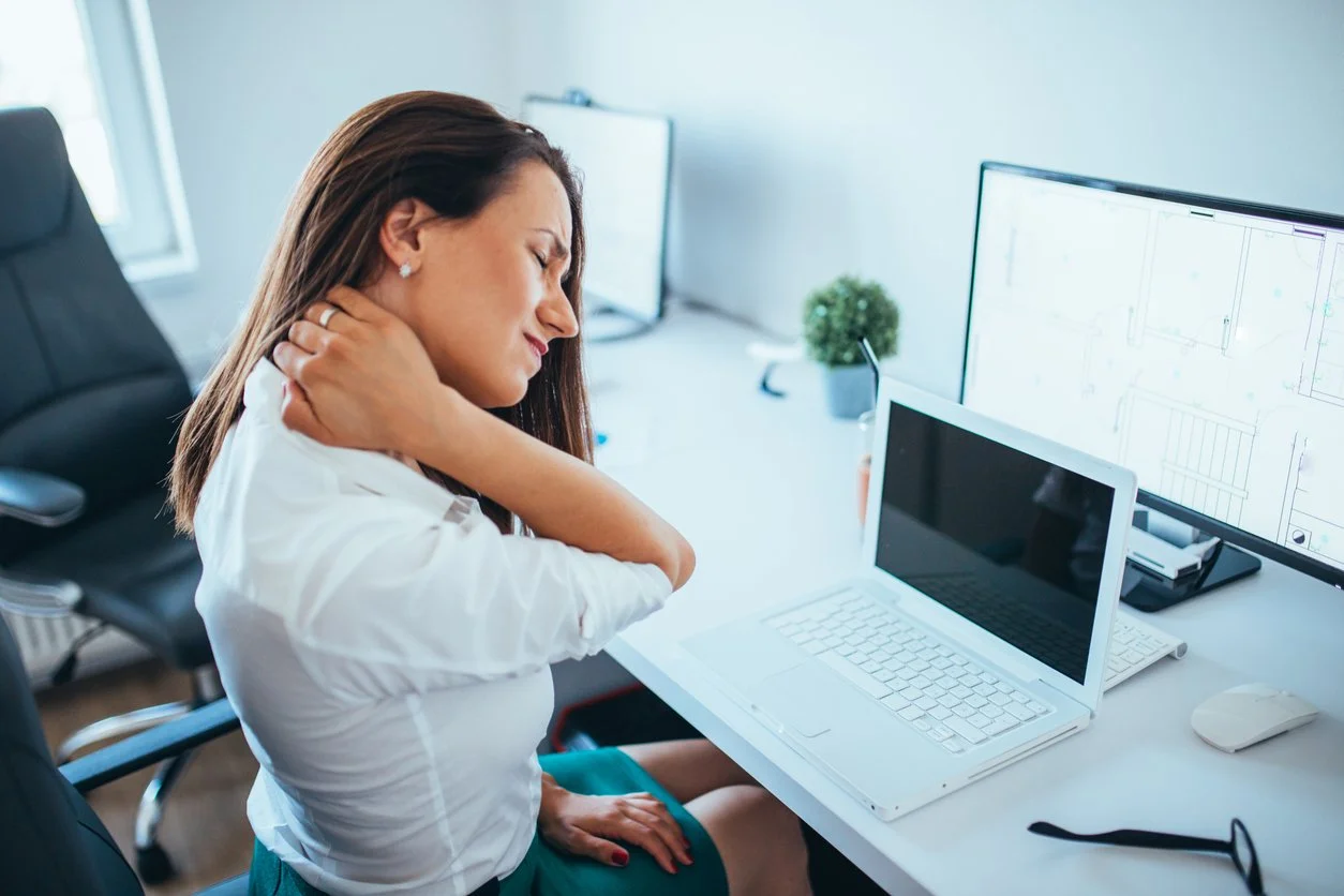 Woman sitting at a desk with her eyes closed, neck pain relief, surrounded by a computer, laptop, and office supplies in a bright office